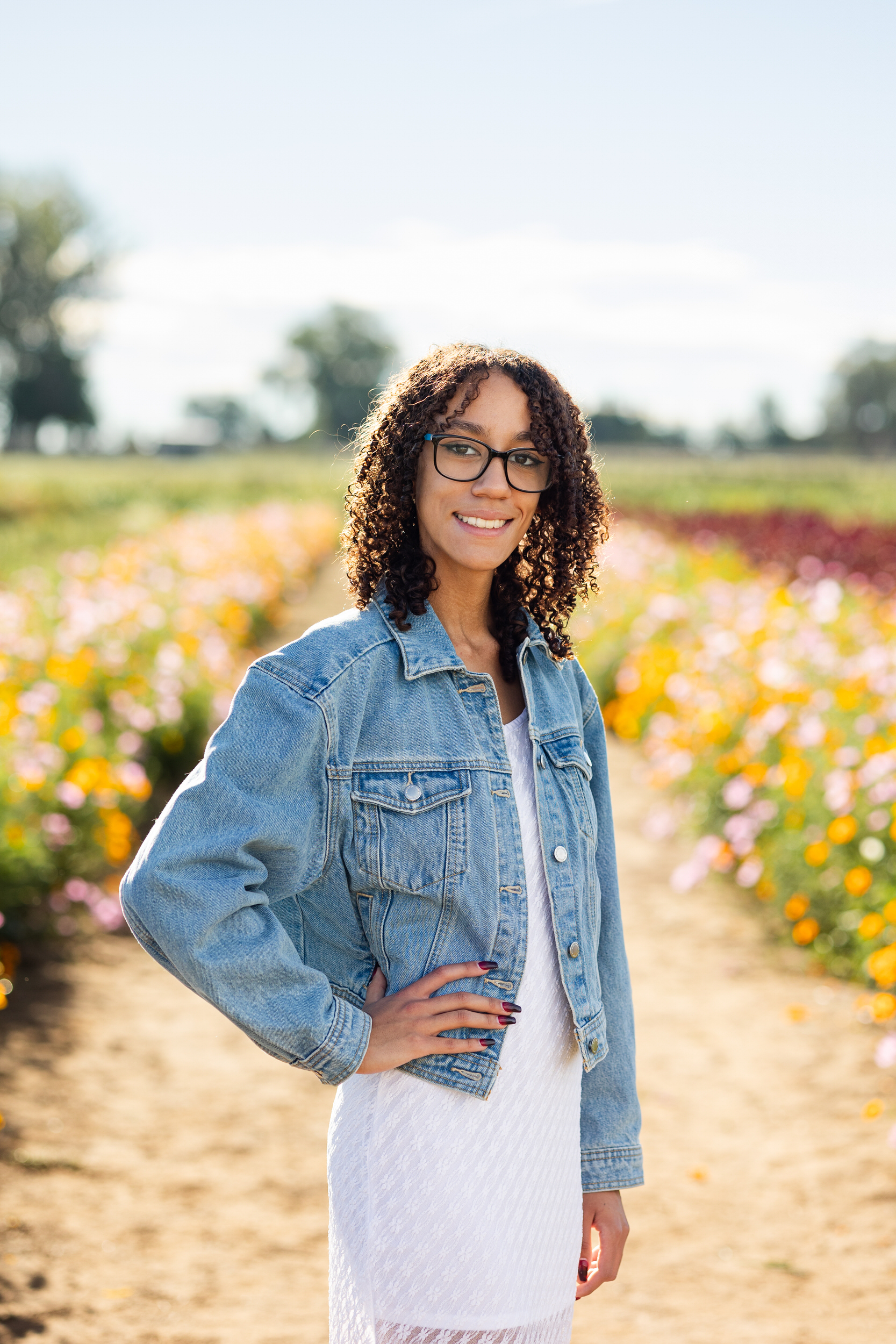 A young woman in a white dress and denim jacket stands in a field of flowers with one hand on her hip and smiles at the camera.