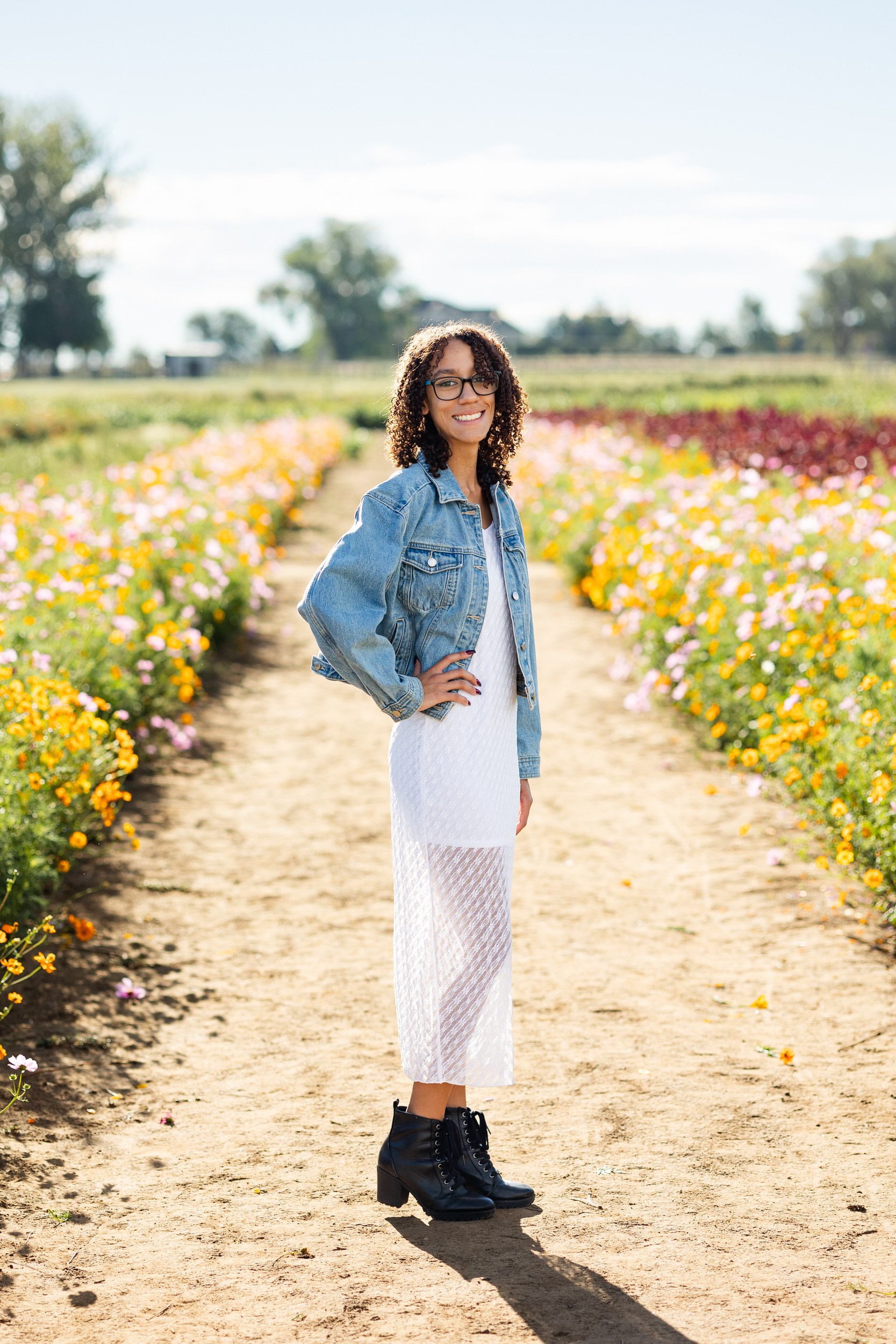 A young woman in a white dress and denim jacket stands in a field of flowers with one hand on her hip and smiles at the camera.