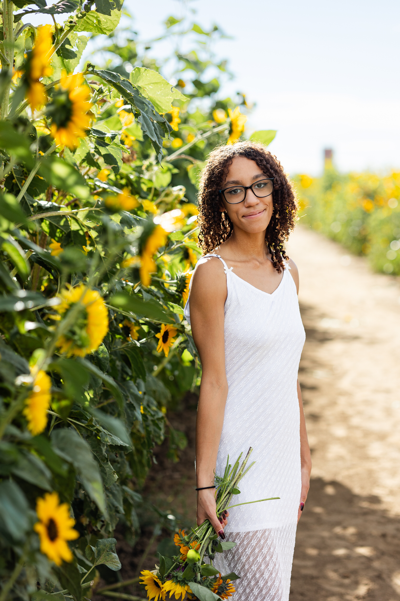 A young woman in a white dress stands in a field of sunflowers and smiles at the camera.