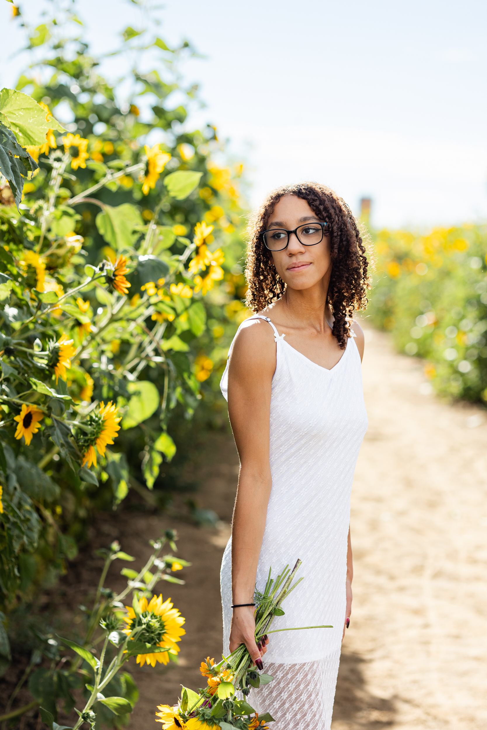 A young woman in a white dress stands in a field of sunflowers and looks over her shoulder.
