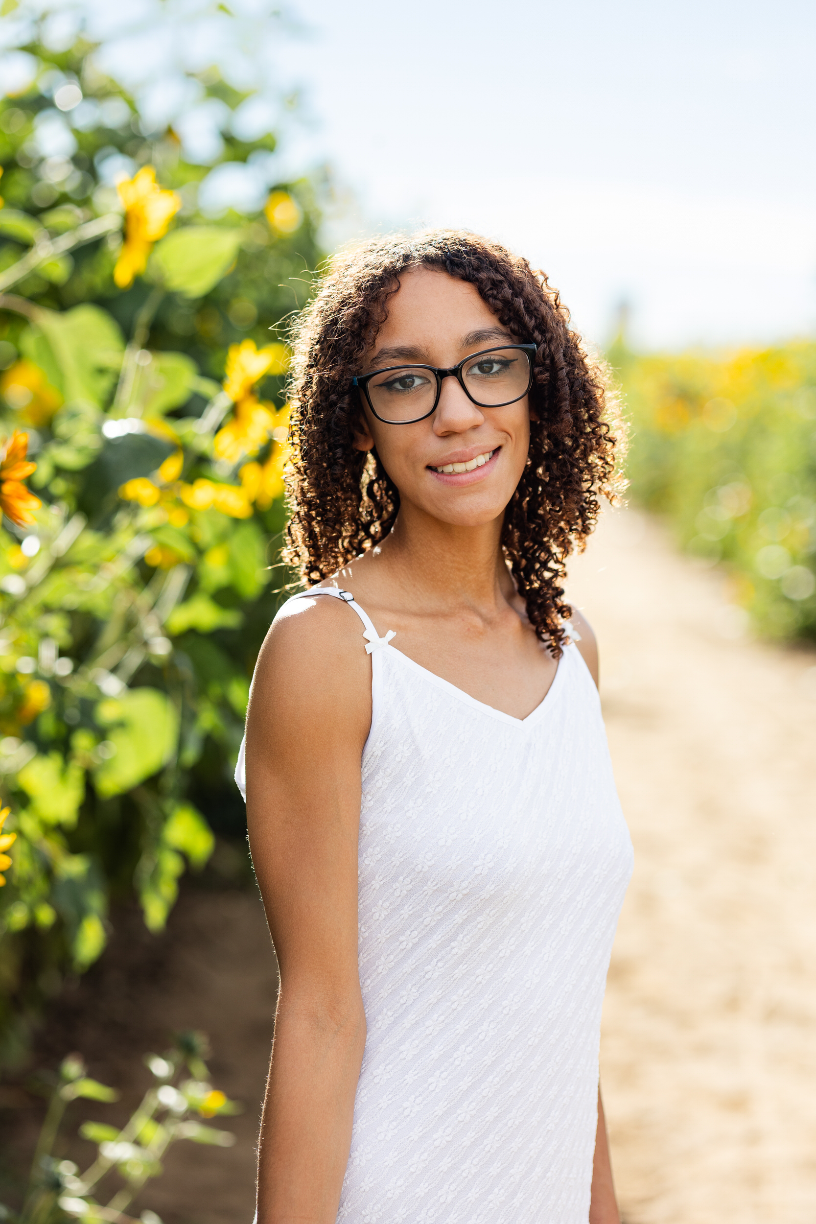 A young woman in a white dress stands in a sunflower field and smiles at the camera.