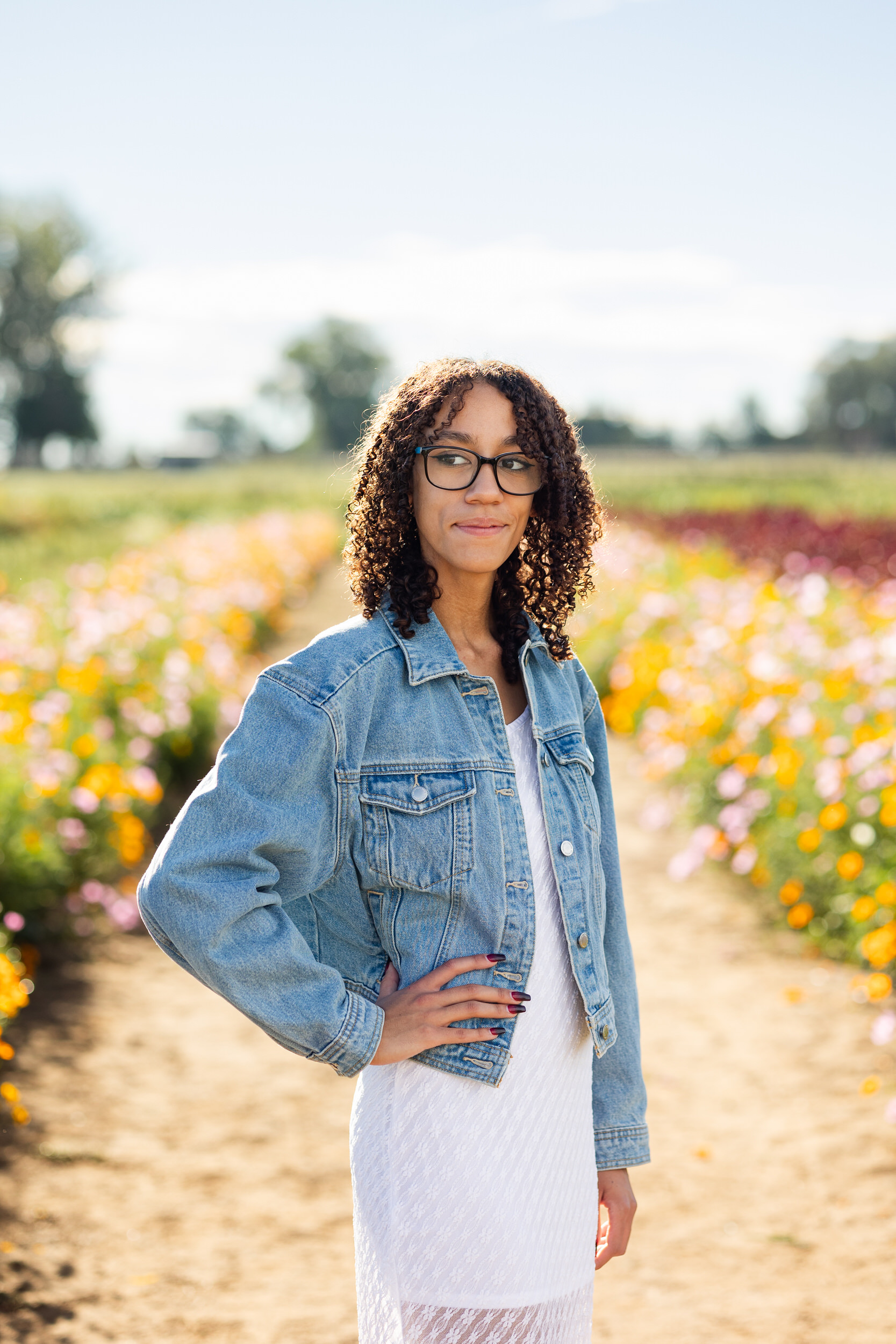 A young woman in a white dress and denim jacket stands in a field of flowers and looks over her shoulder.