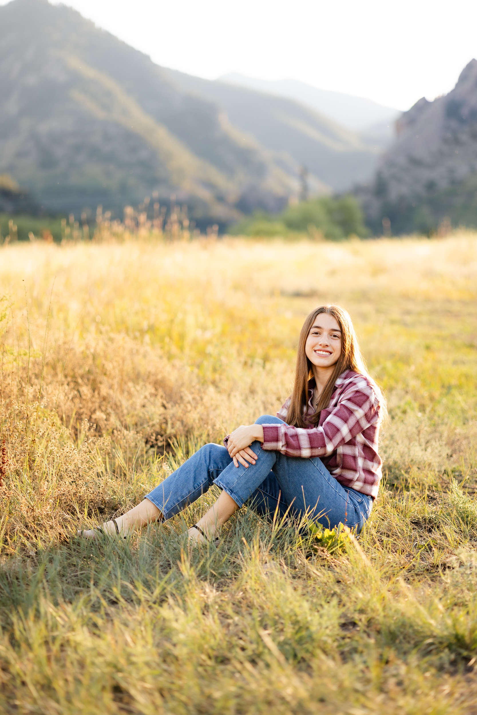 A young woman in a red flannel and blue jeans sits in a field with mountains in the background and smiles at the camera.