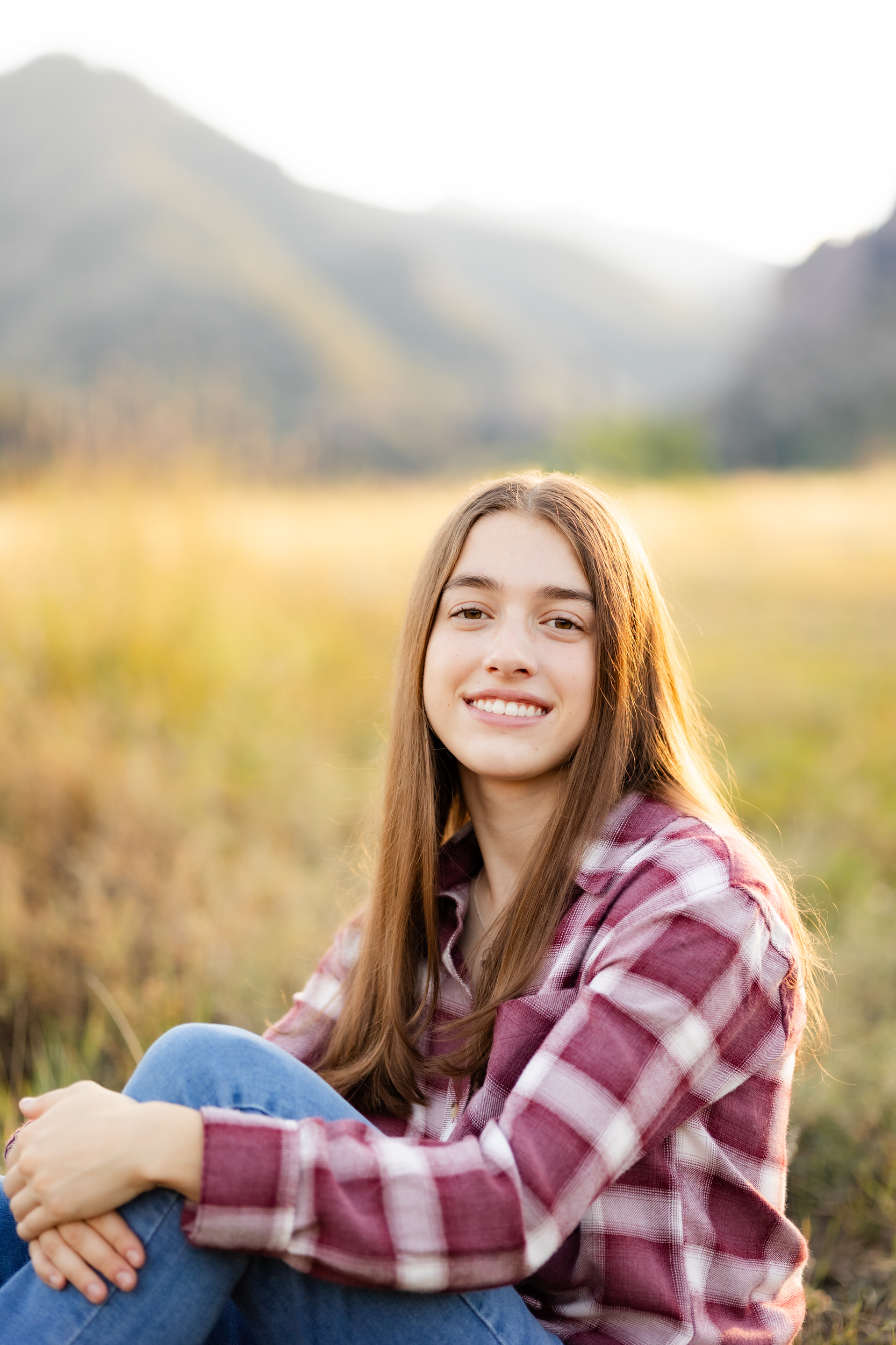A young woman in a red flannel and blue jeans sits in a field with mountains in the background and smiles at the camera.
