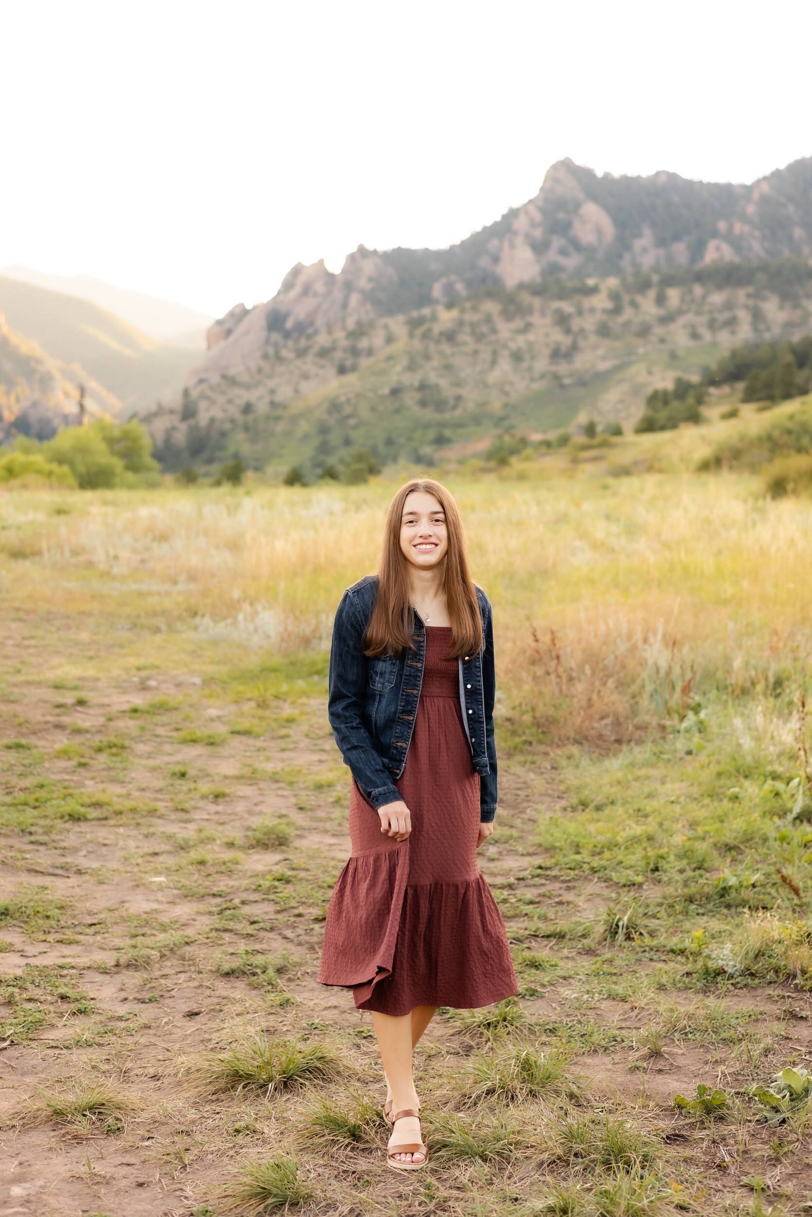 A young woman in a burgundy dress and denim jacket walks through a field with mountains in the background and smiles at the camera.