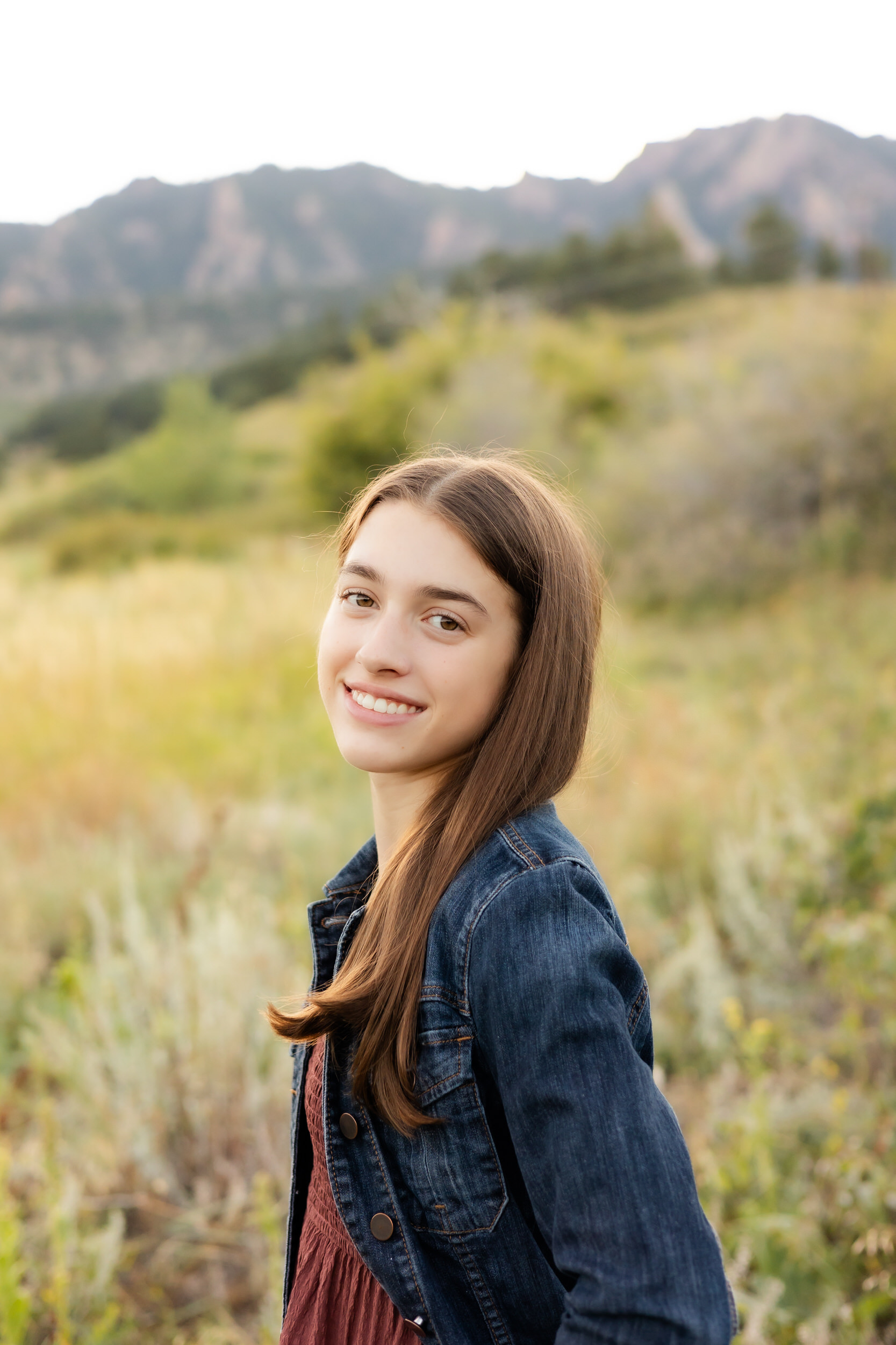 A young woman in burgundy dress and denim jacket smiles over her shoulder at the camera.