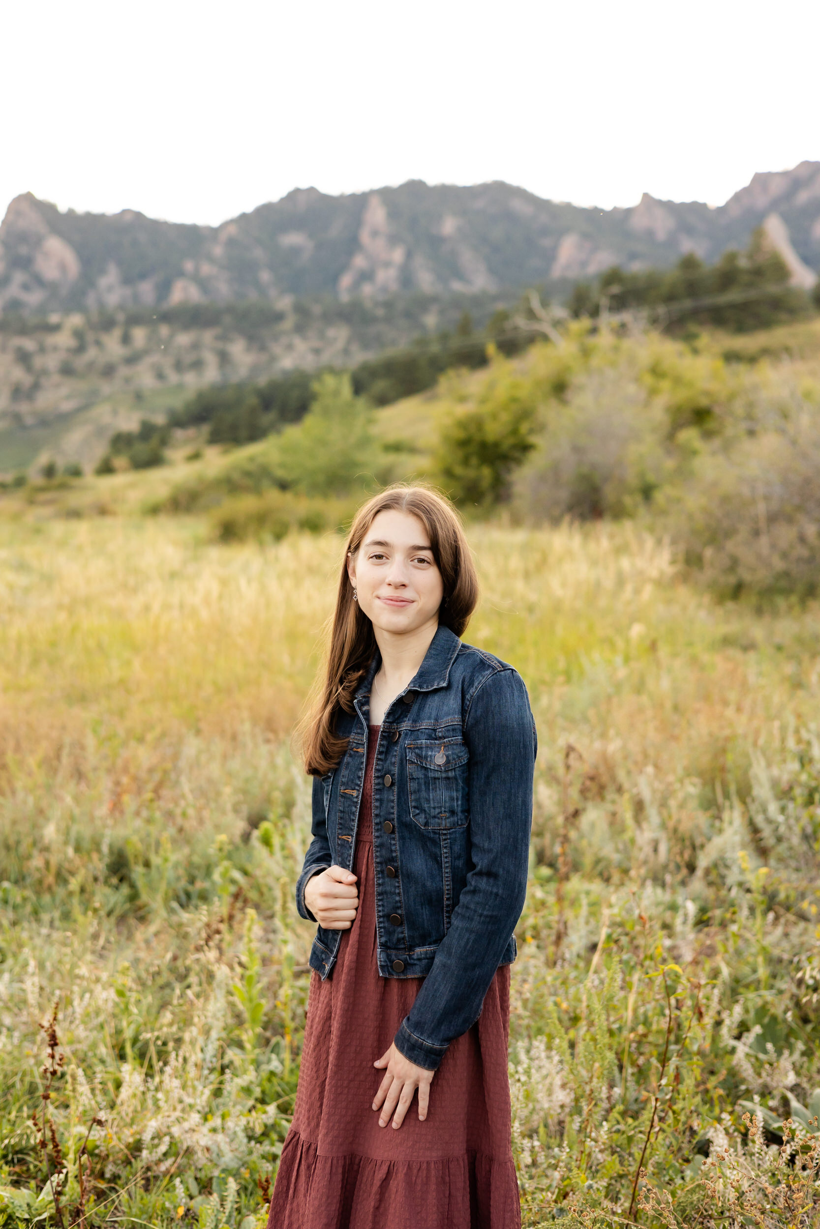 A young woman in a burgundy dress and denim jacket stands in a field with mountains in the background and smiles at the camera.