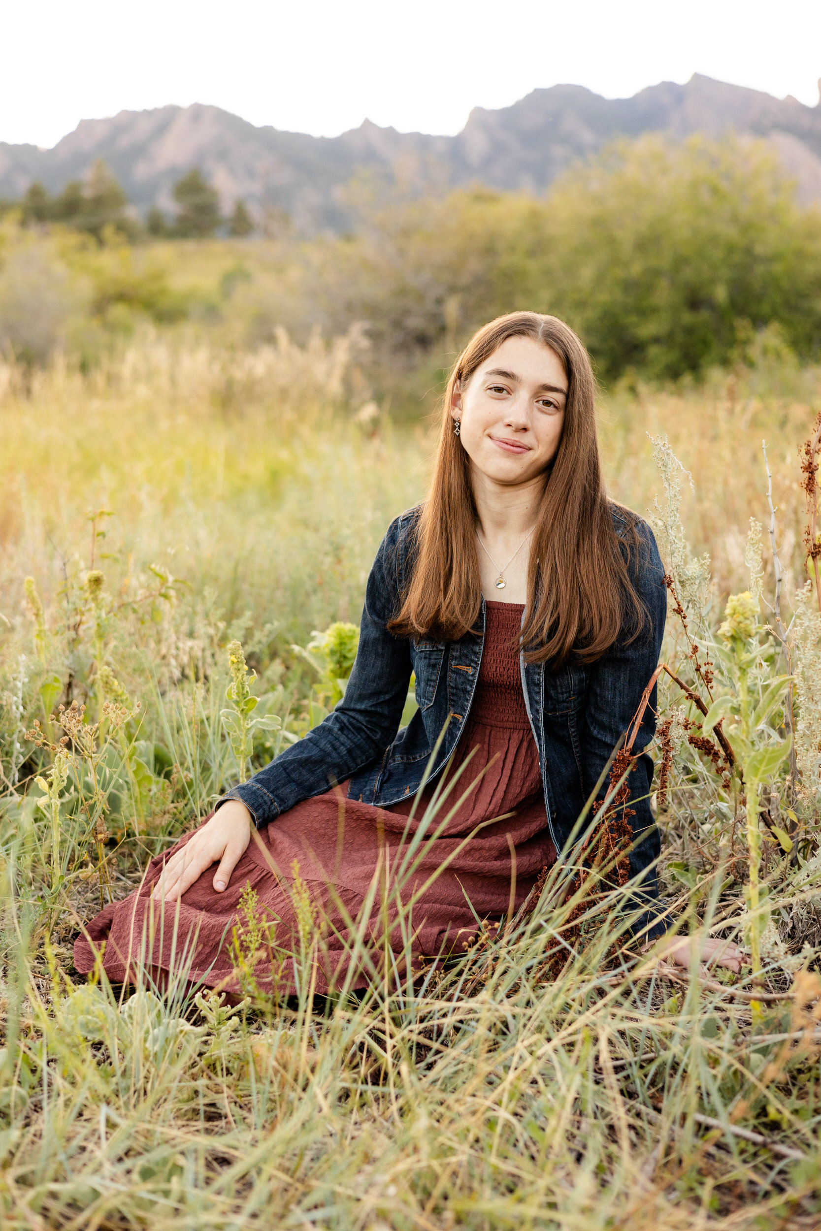 A young woman in a burgundy dress and denim jacket sits in a field with mountains in the background and smiles at the camera.