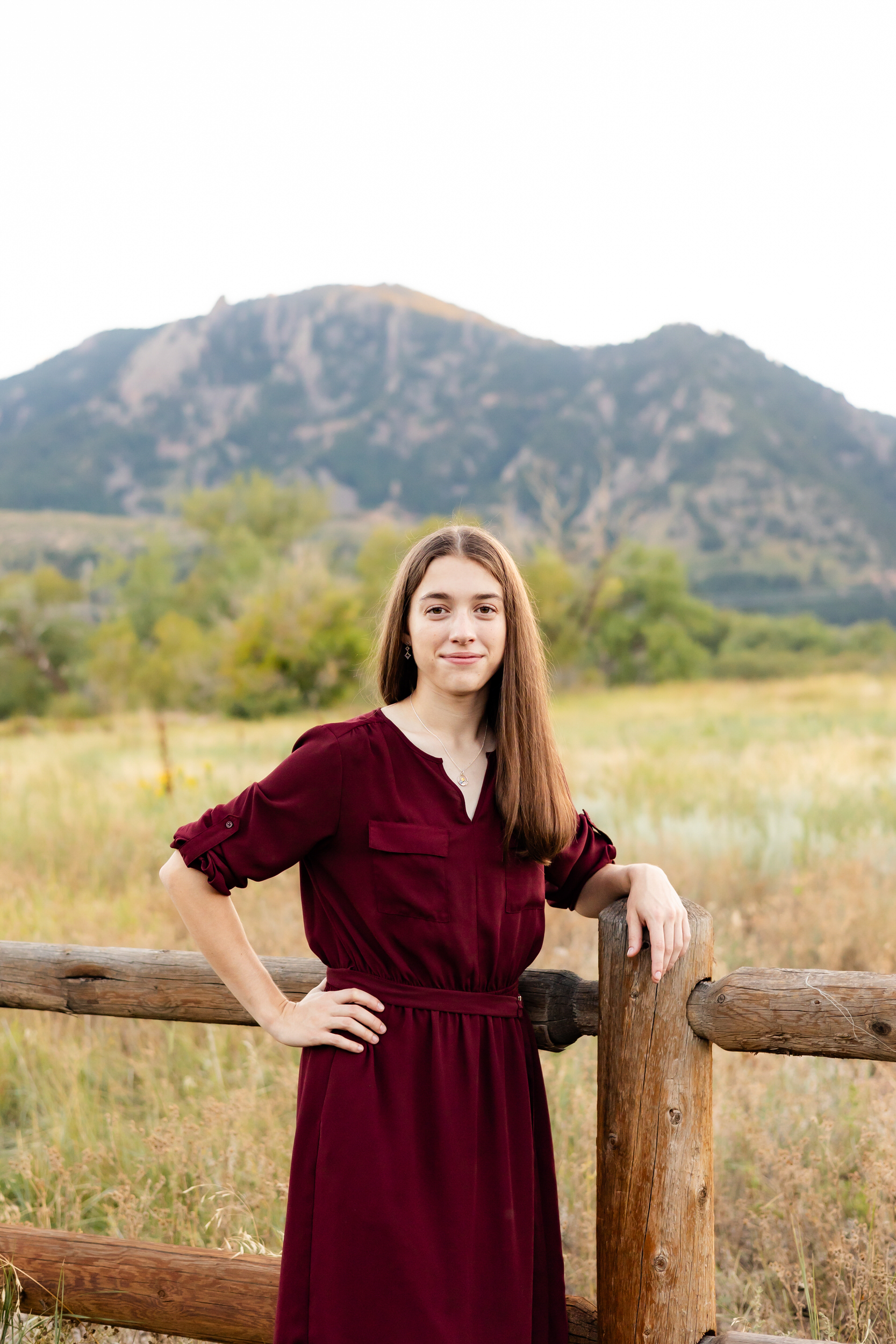A young woman in a burgundy dress leans against a trailside fence with mountains in the background and smiles at the camera.