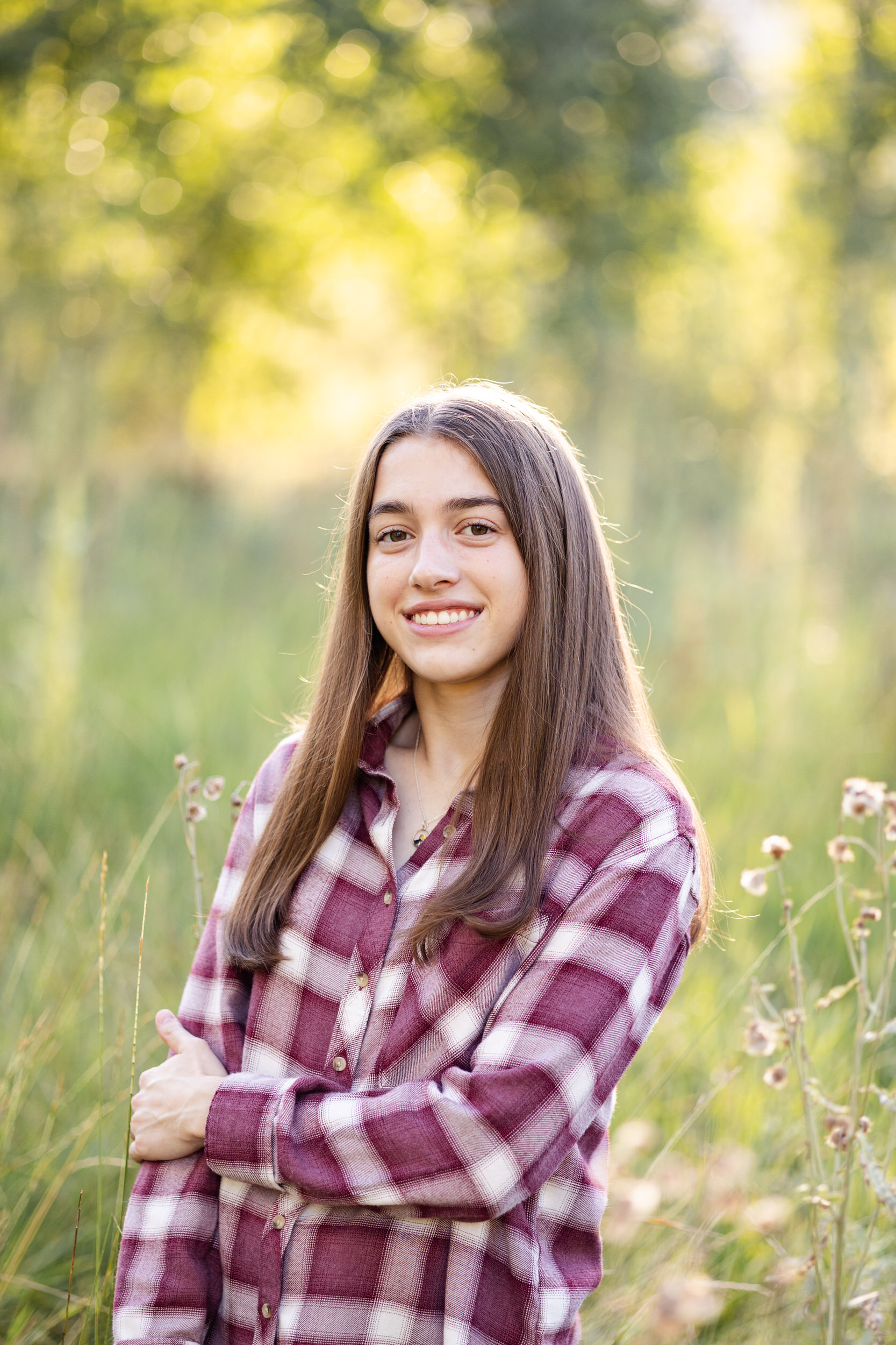 A young woman in a red flannel stands in a field and smiles at the camera.