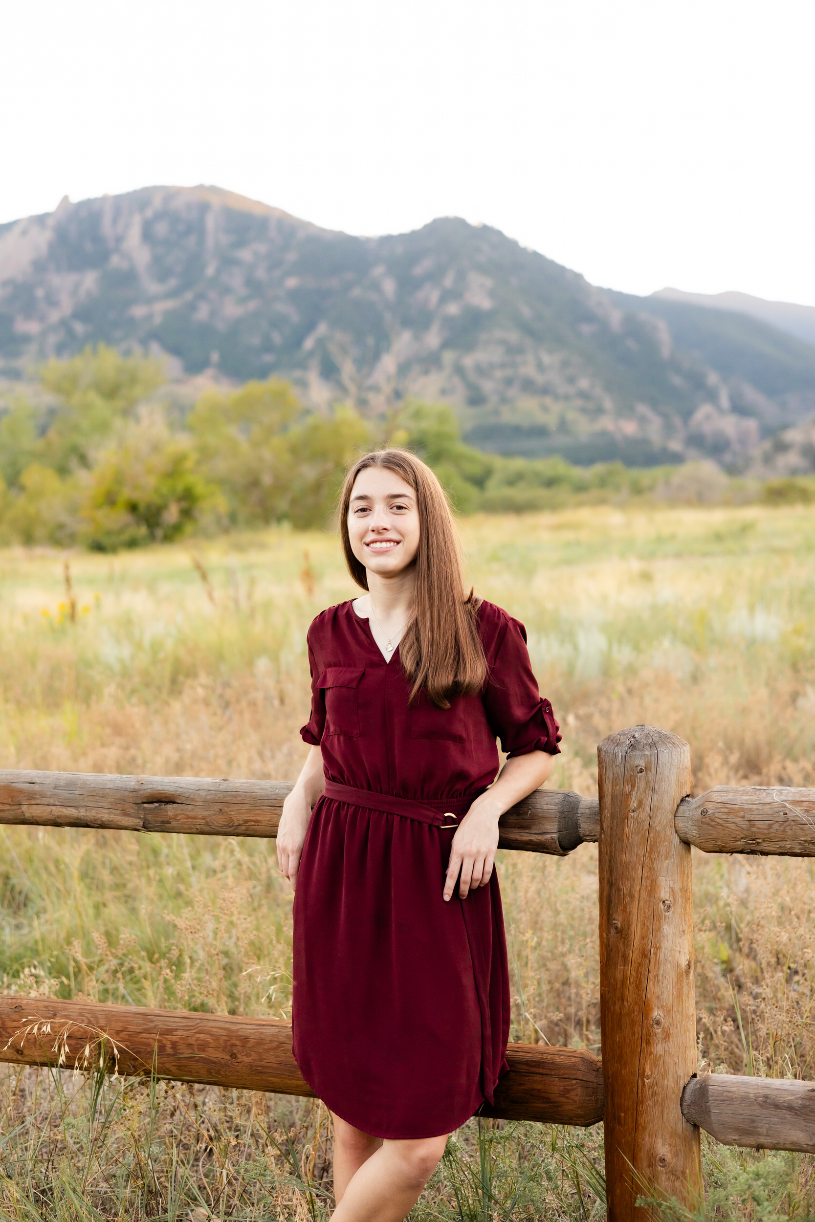 A young woman in a burgundy dress leans against a trailside fence with mountains in the background and smiles at the camera.