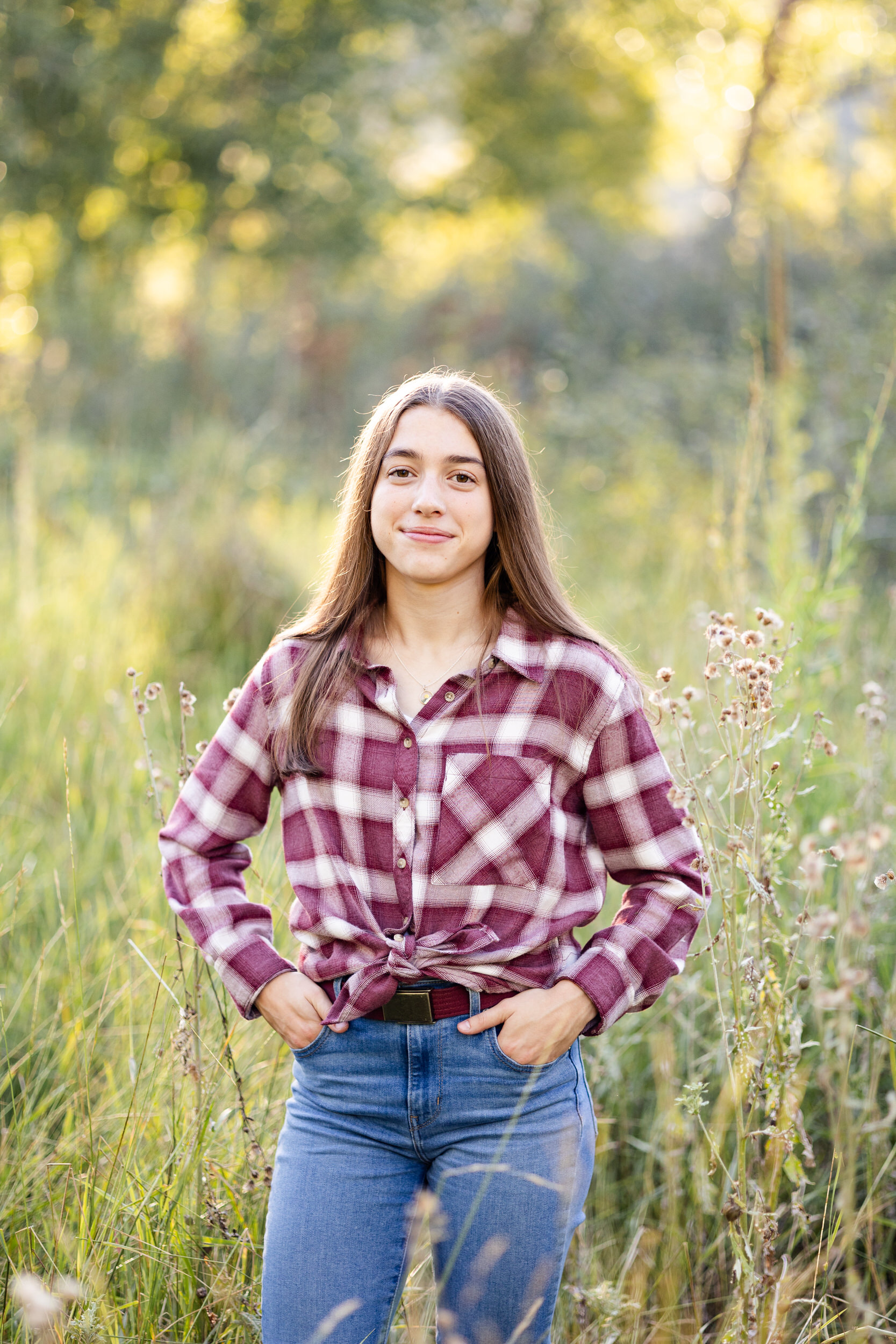 A young woman in a red flannel stands in a field and smiles at the camera.
