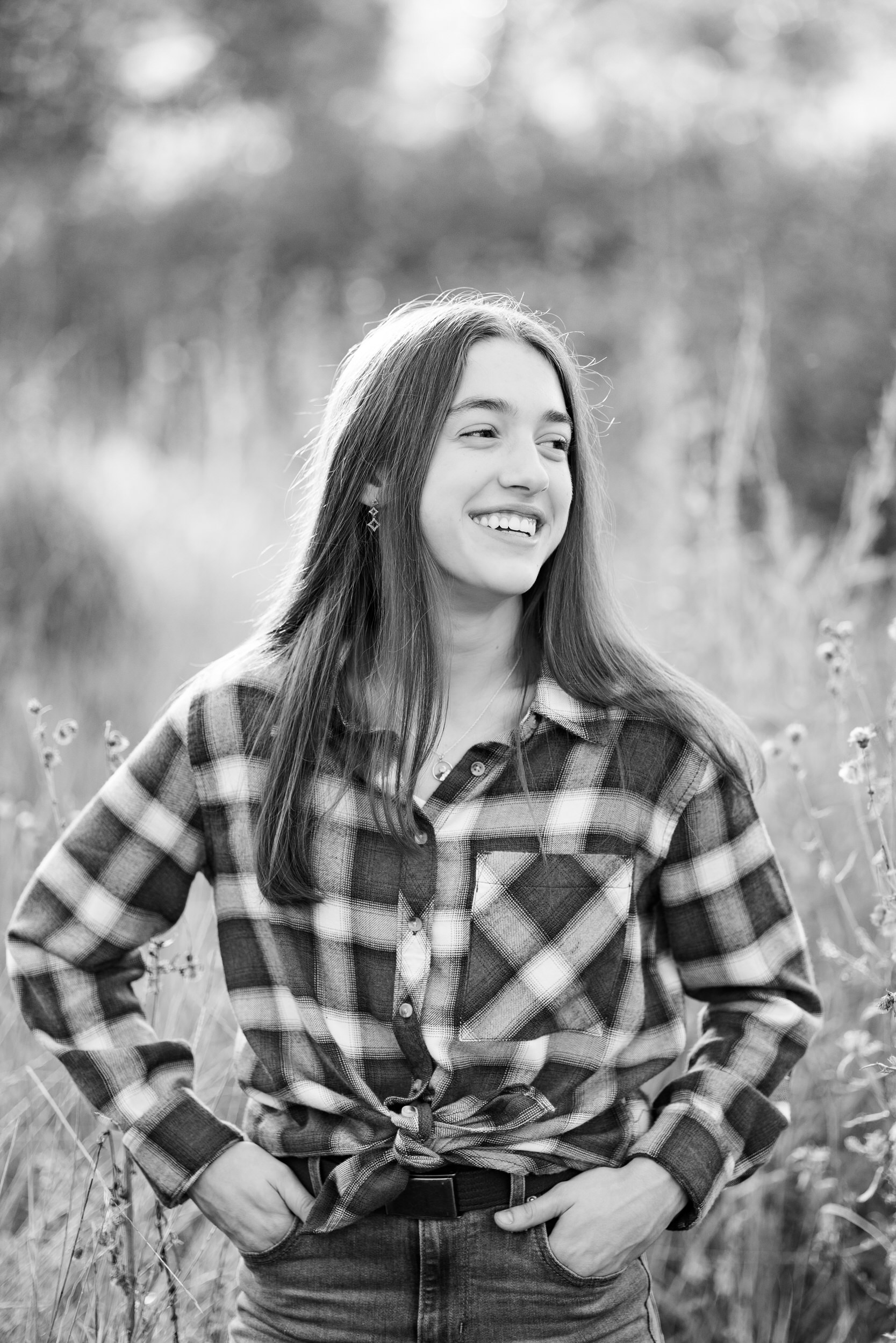 A young woman in a red flannel stands in a field and smiles over her shoulder off-camera.