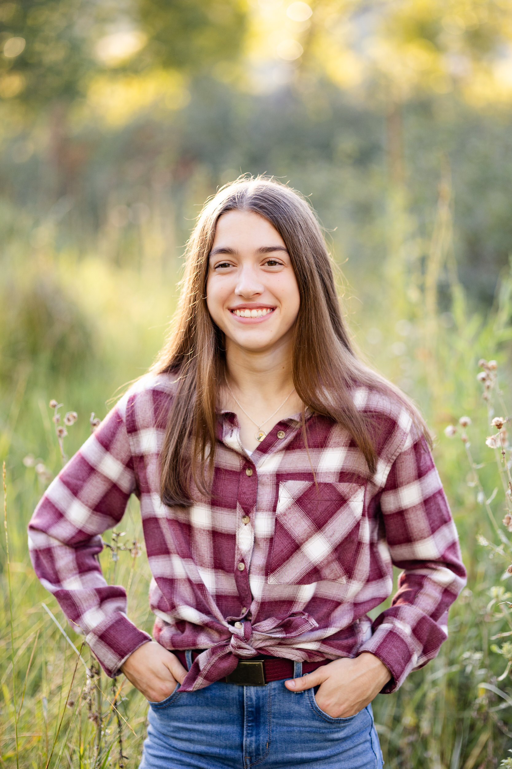 A young woman in a red flannel stands in a field and smiles at the camera.