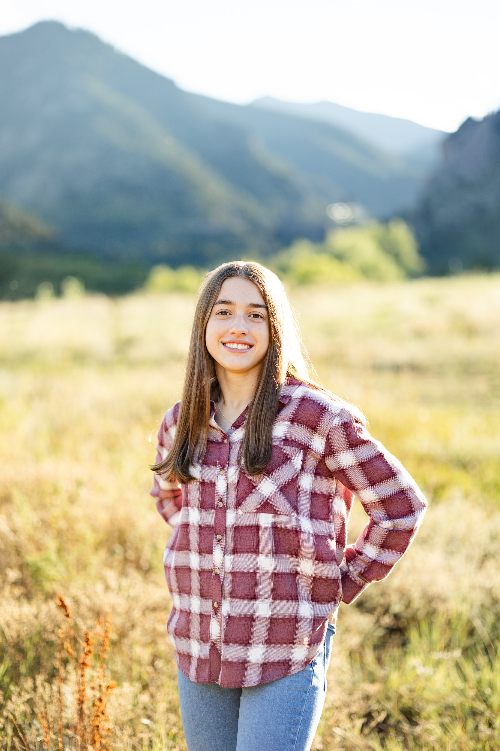 A young woman in a red flannel and blue jeans smiles at the camera in a field with mountains in the background.