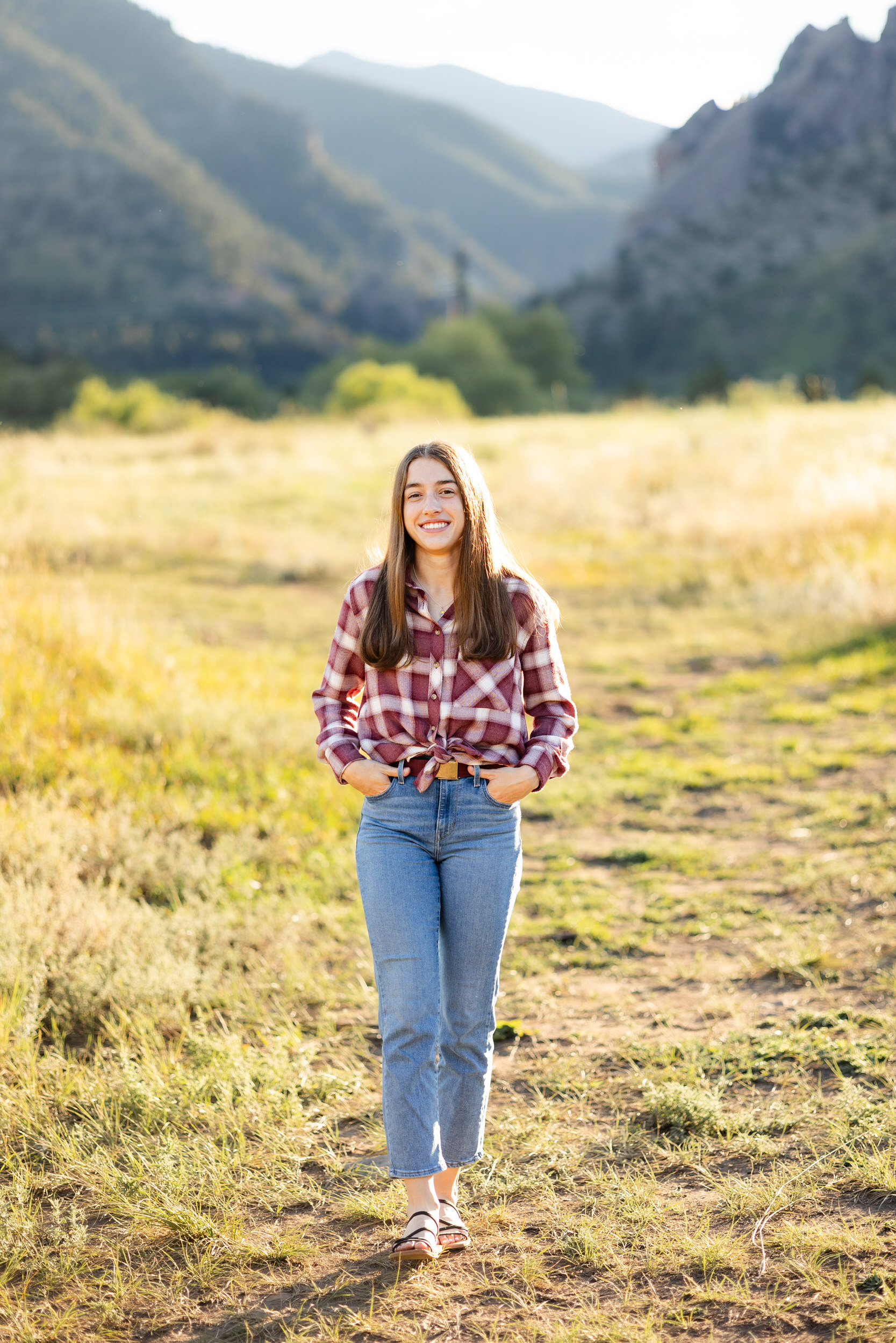 A young woman in a red flannel and blue jeans walks through a field and smiles at the camera with mountains in the background.