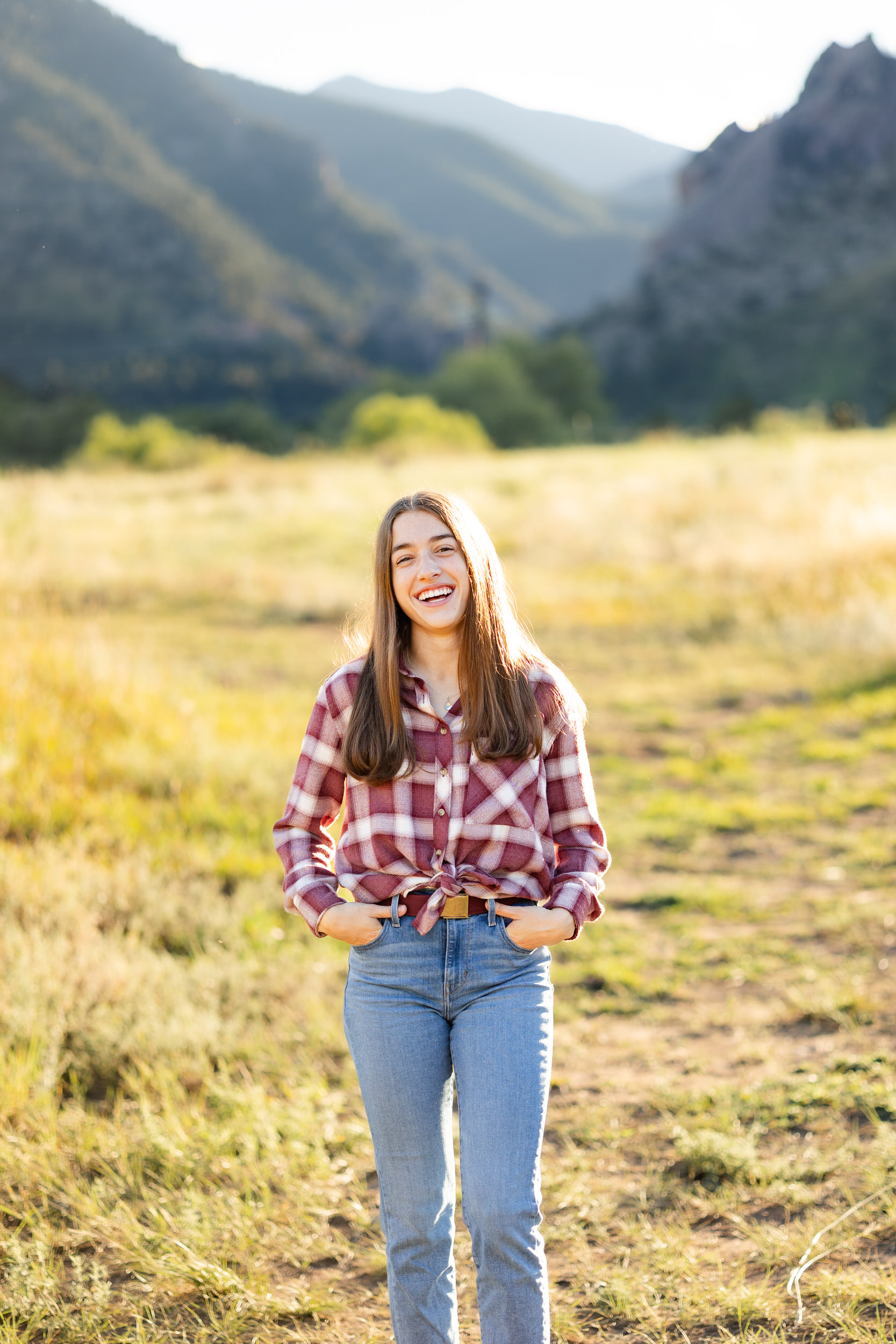 A young woman in a red flannel and blue jeans walks through a field and laughs at the camera with mountains in the background.