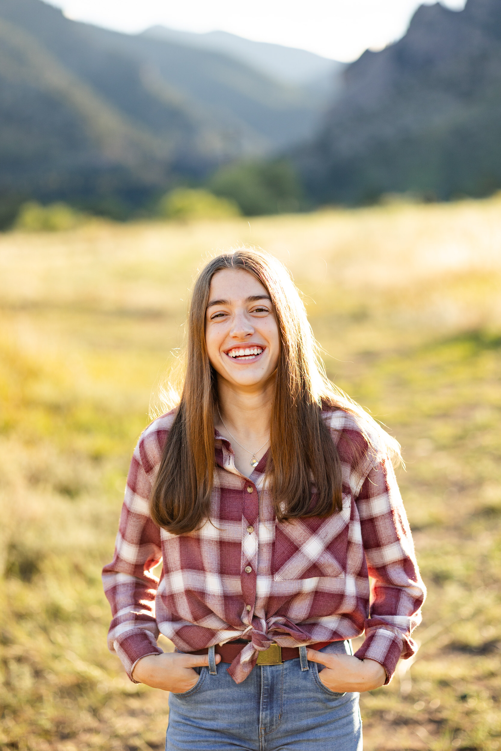 A young woman in a red flannel and blue jeans smiles at the camera with mountains in the background.