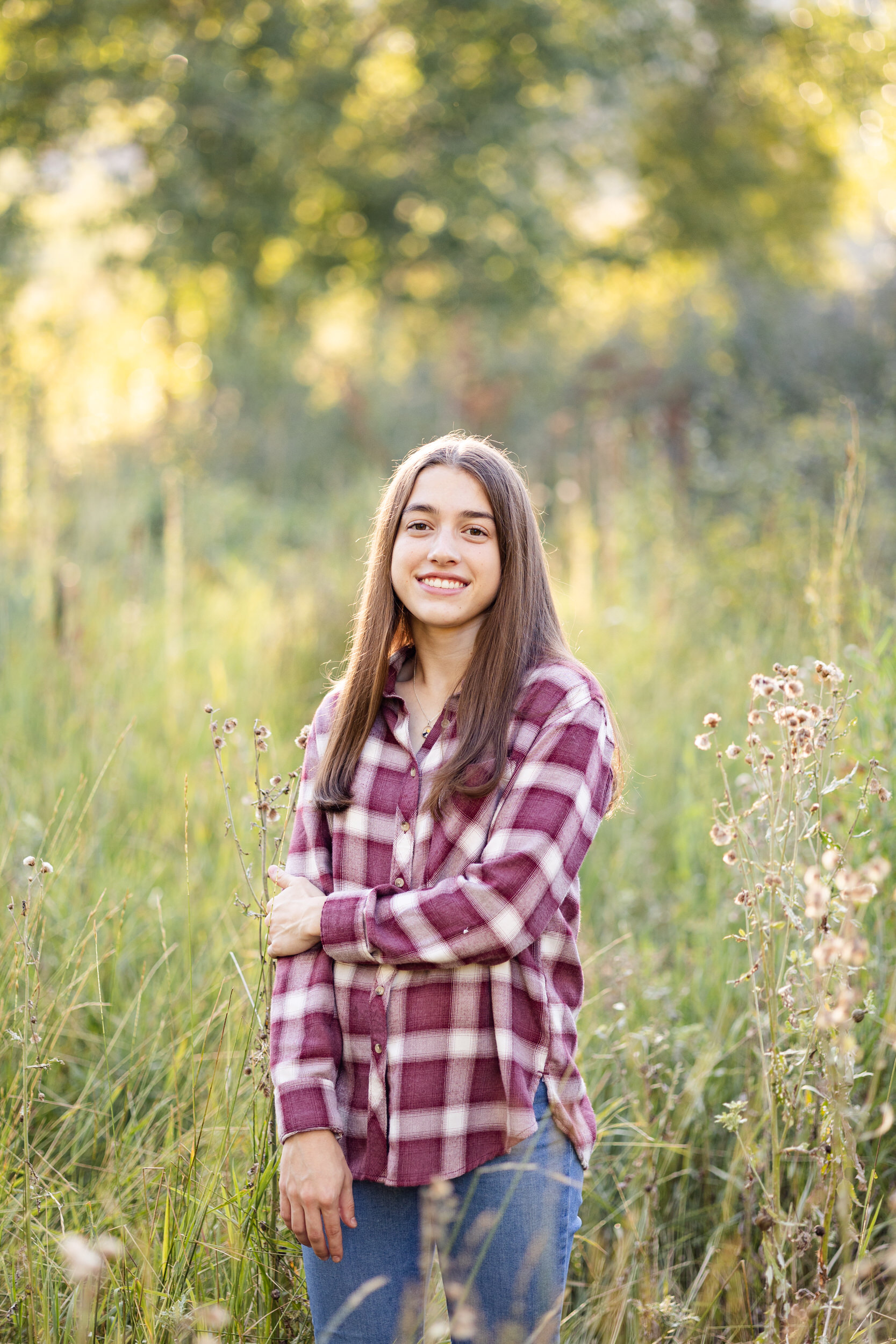 A young woman in a red flannel shirt and blue jeans stands in a field and smiles at the camera.