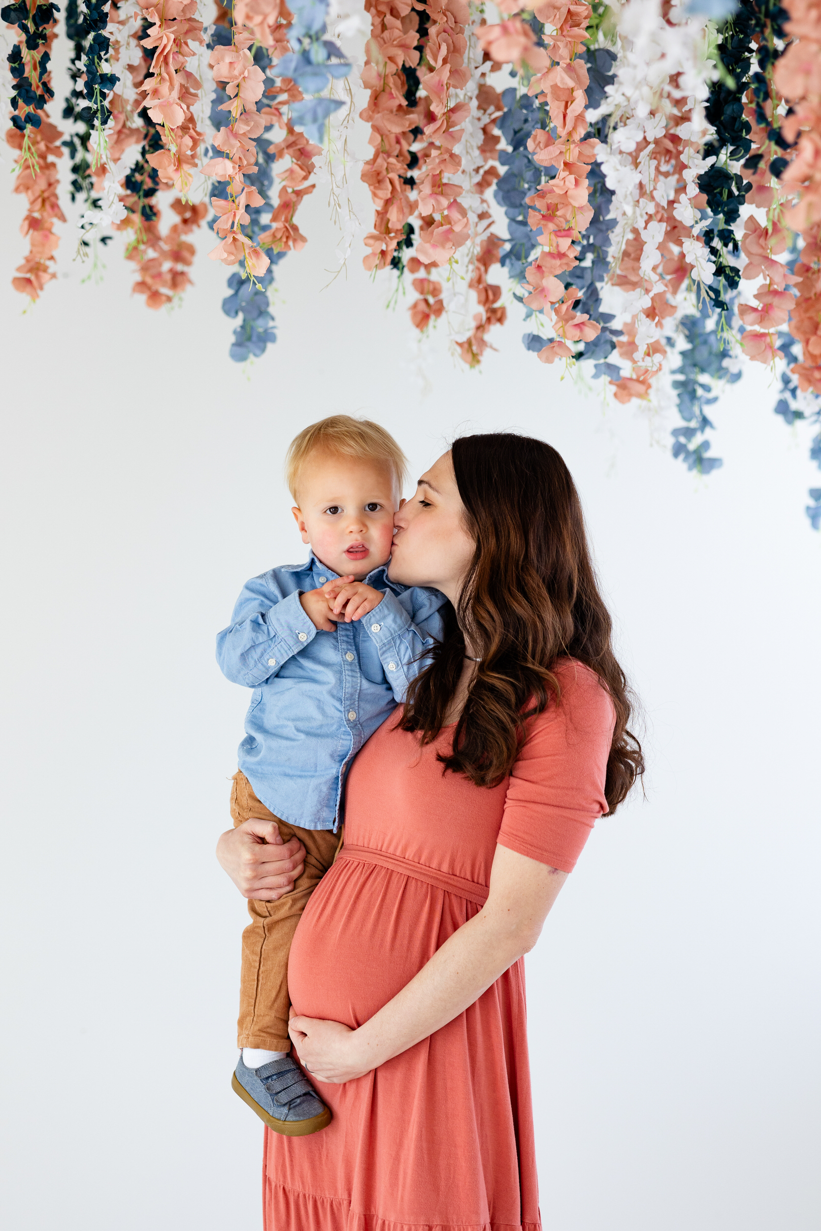 A mom holds her toddler son in one hand and kisses her cheek and holds her pregnant belly with the other hand.