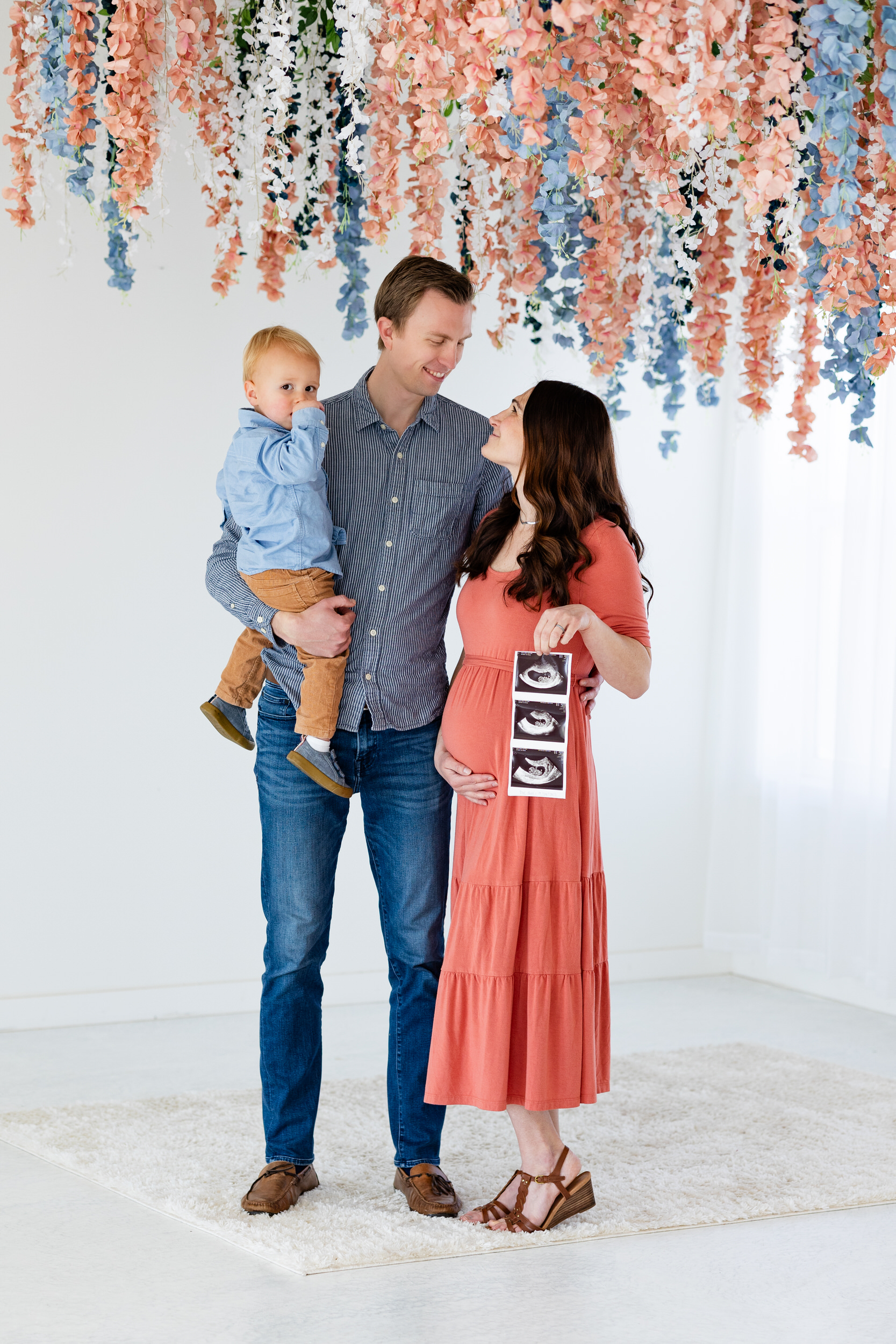A mom and dad smile at each other while holding their toddler son and ultrasound photos.