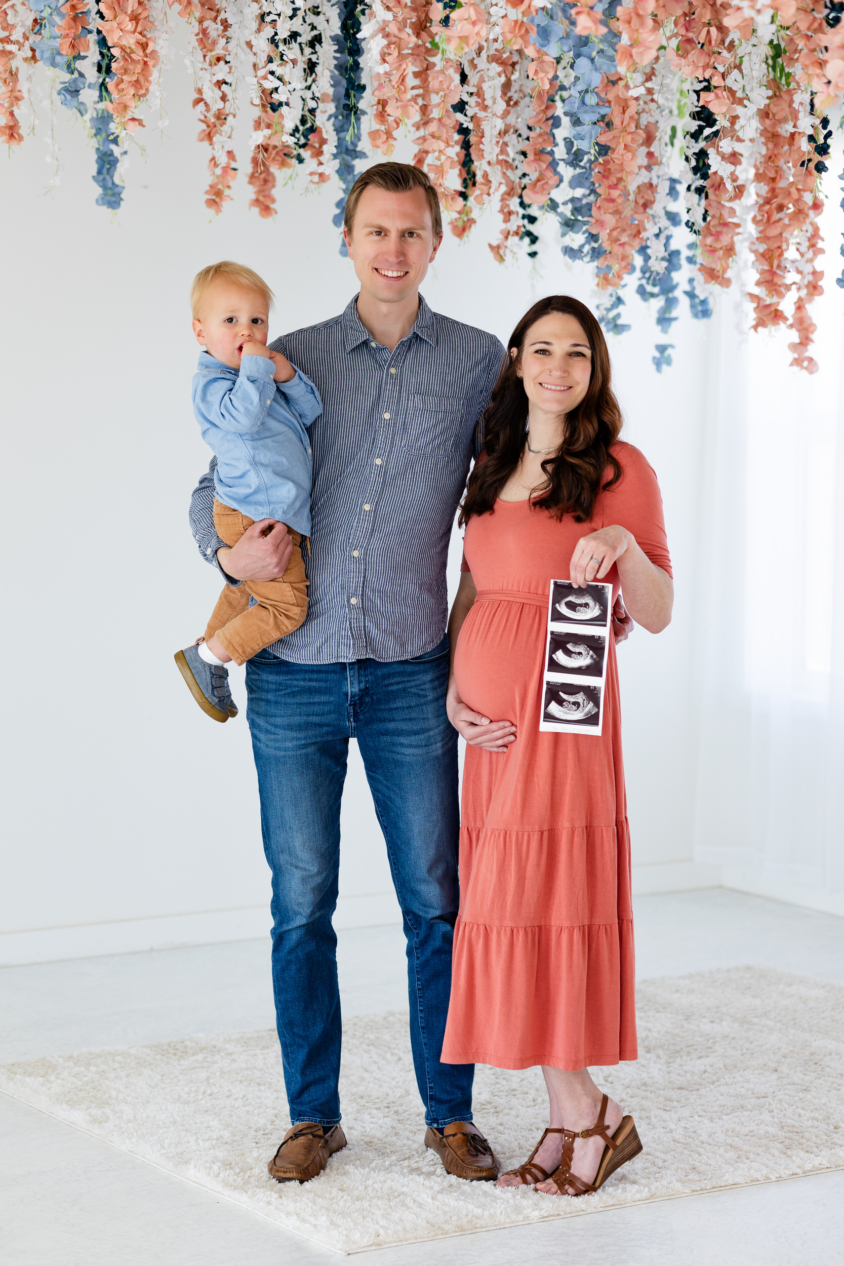 A mom and dad smile at the camera while holding their toddler son and ultrasound photos.