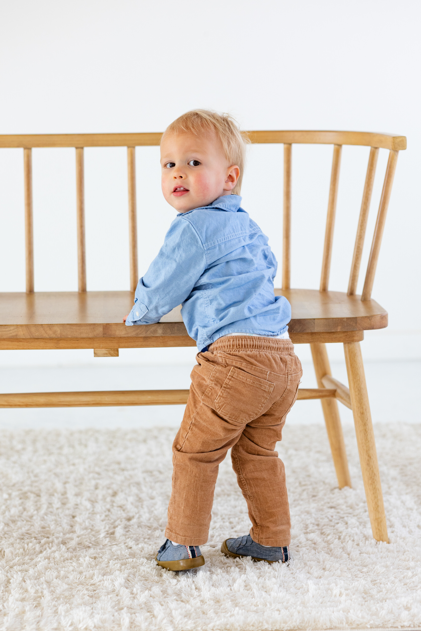 A toddler boy stands in front of a bench and looks back over his shoulder at the camera.