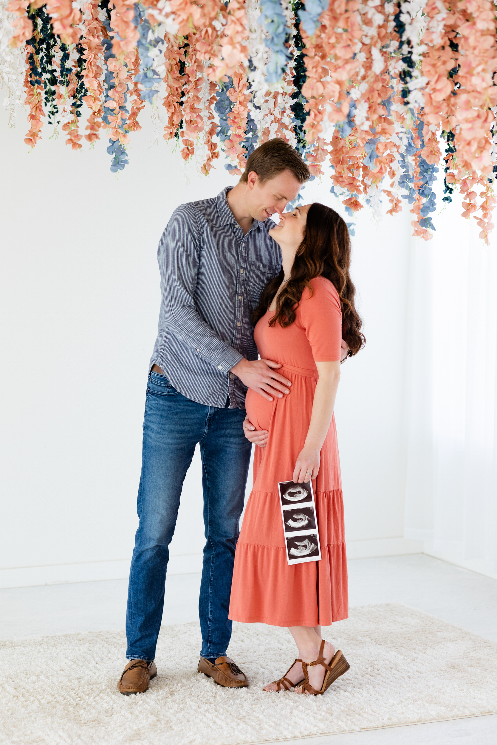 A couple stands nose to nose as they smile and the woman holds an ultrasound picture and she and her husband both hold the baby bump.