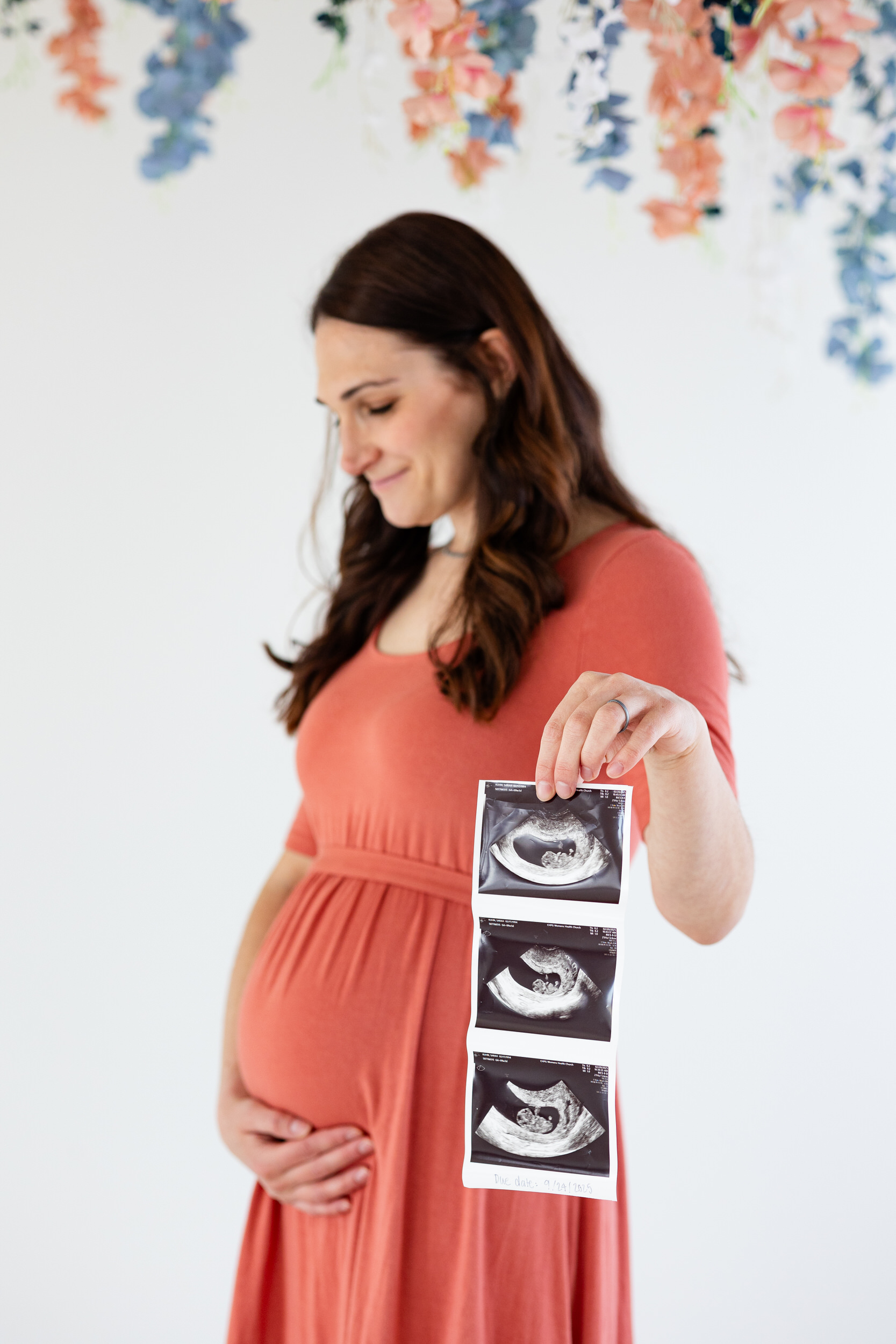 A woman smiles at her pregnant belly with one hand and ultrasound photos in the other hand.