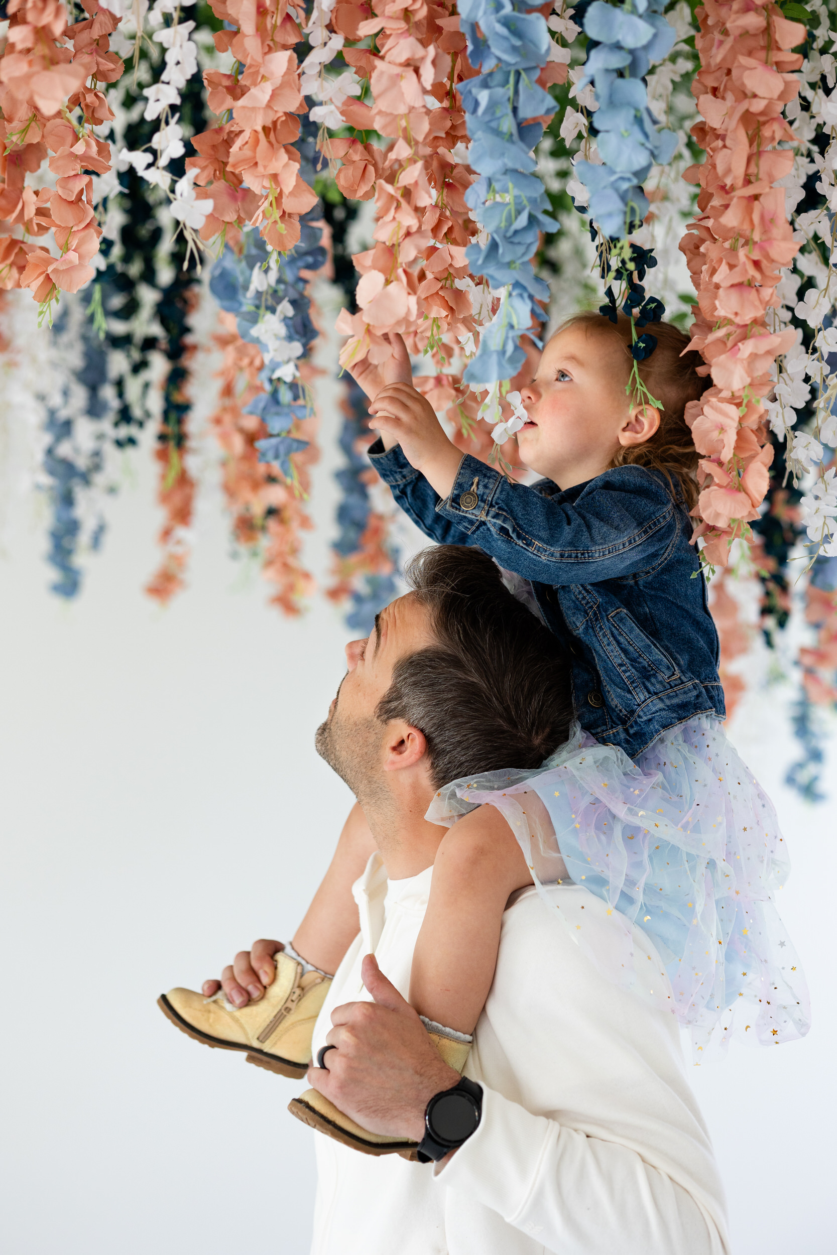 A toddler girl sits on her dad's shoulders and looks at flowers hanging from the ceiling.