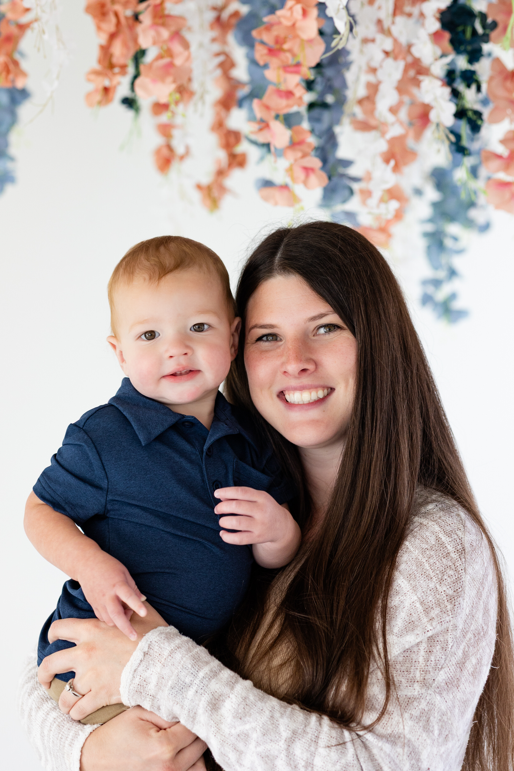 A mom holds her one-year-old son and they both smile at the camera.