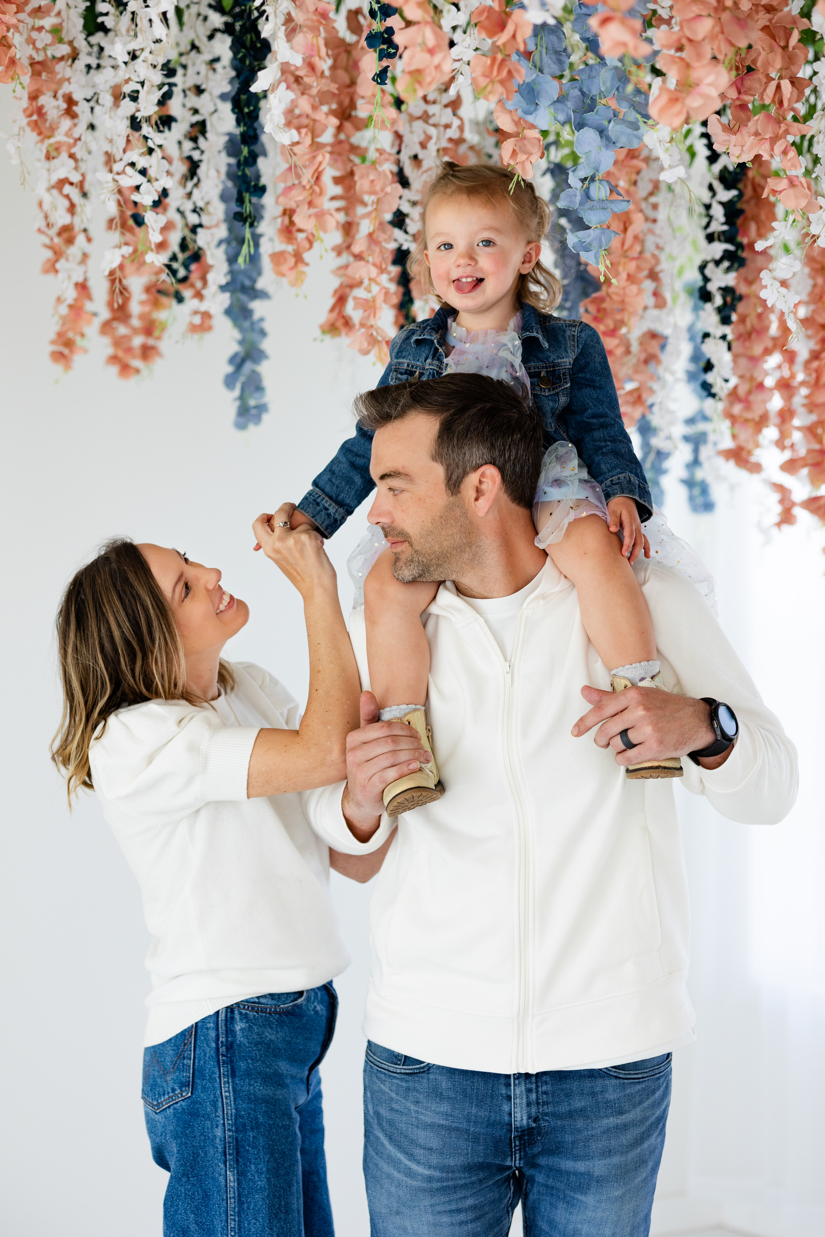 A toddler girl sits on her dad's shoulders while her mom smiles at her and her dad smiles at her mom.