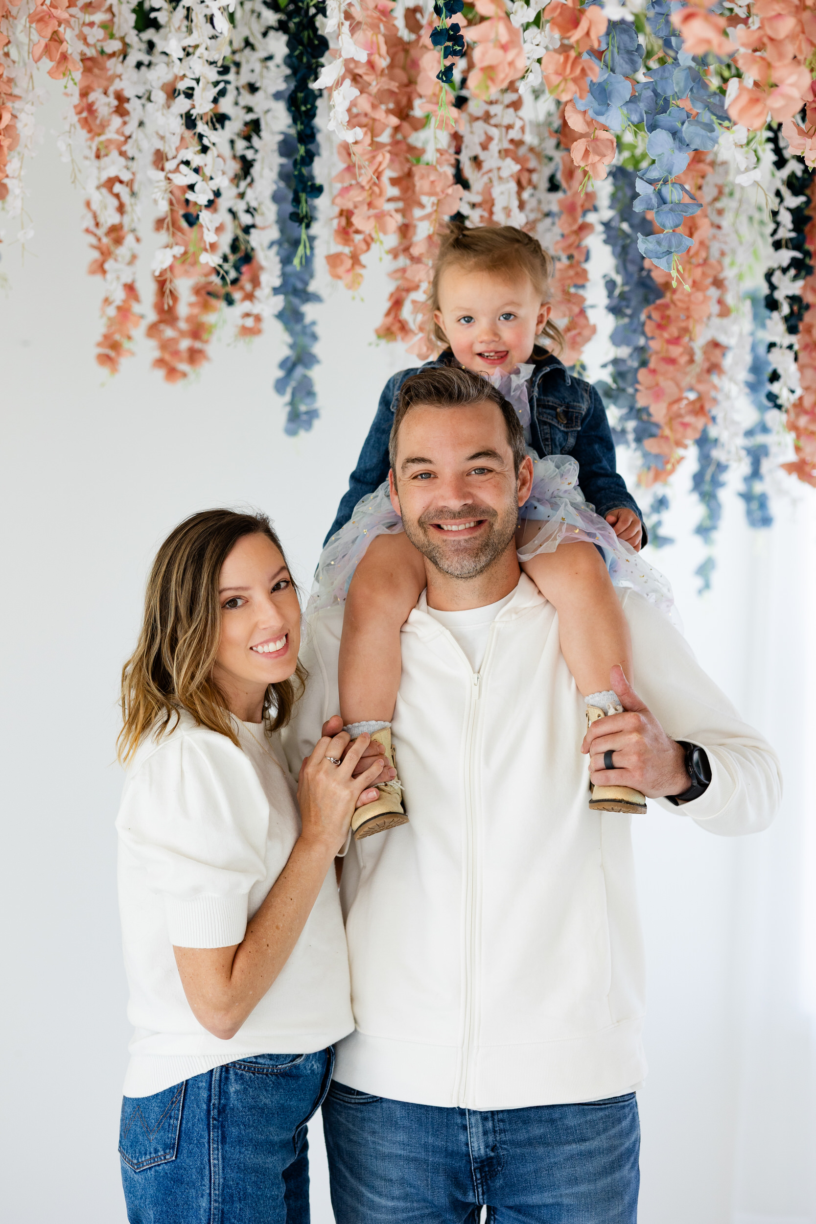 A mom and dad smile at the camera and their toddler daughter sits on her dad's shoulders and also smiles at the camera.