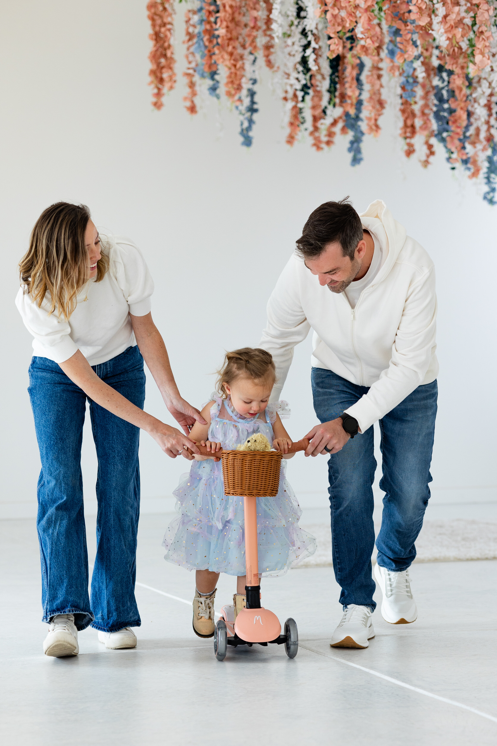 A mom and dad hold the handlebars and run alongside their young daughter on her pink scooter.
