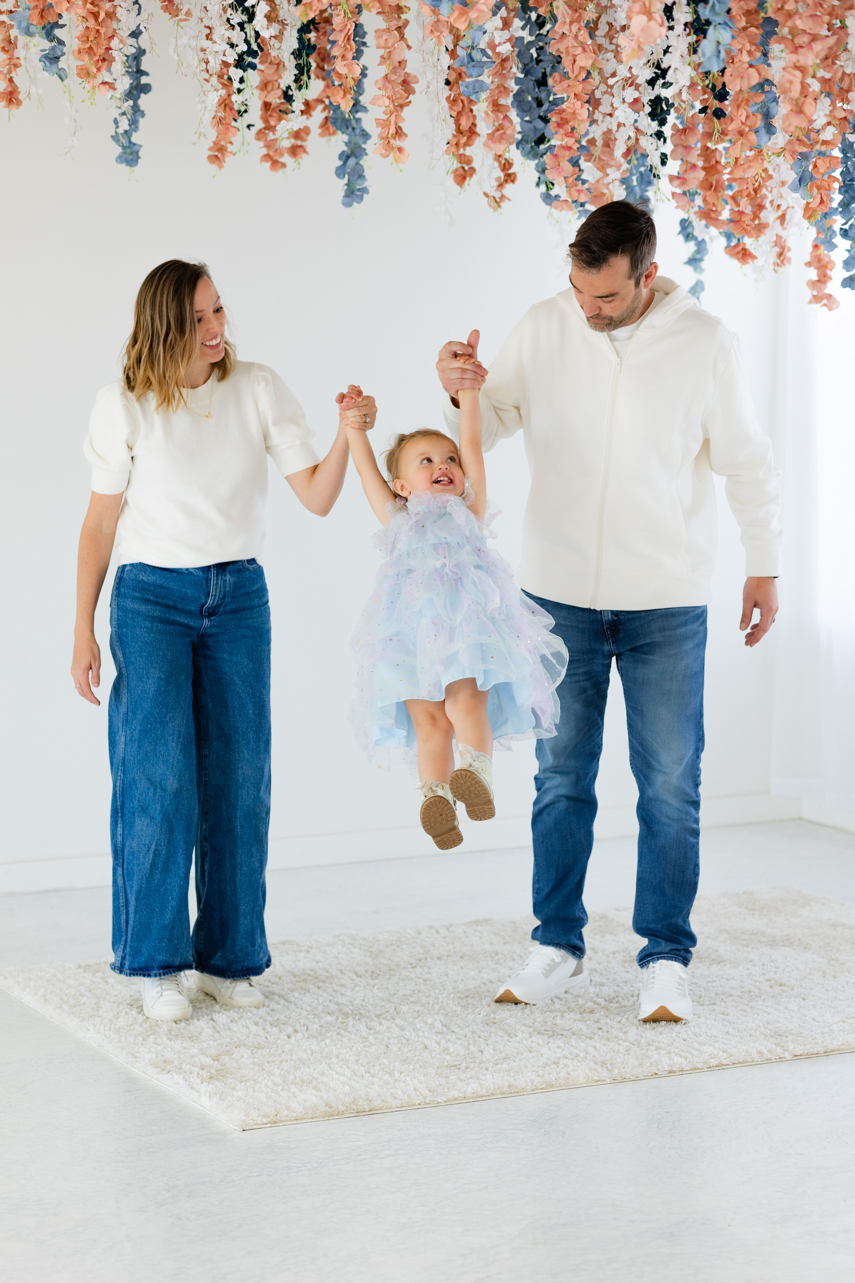 A mom and dad hold their young daughter's hands and swing her between them as she laughs.