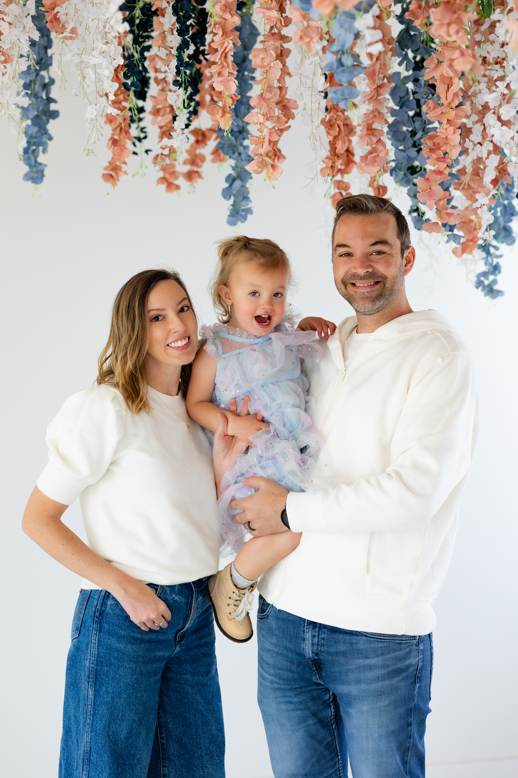 A mom and dad hold their two-year-old daughter between them and they all smile at the camera.
