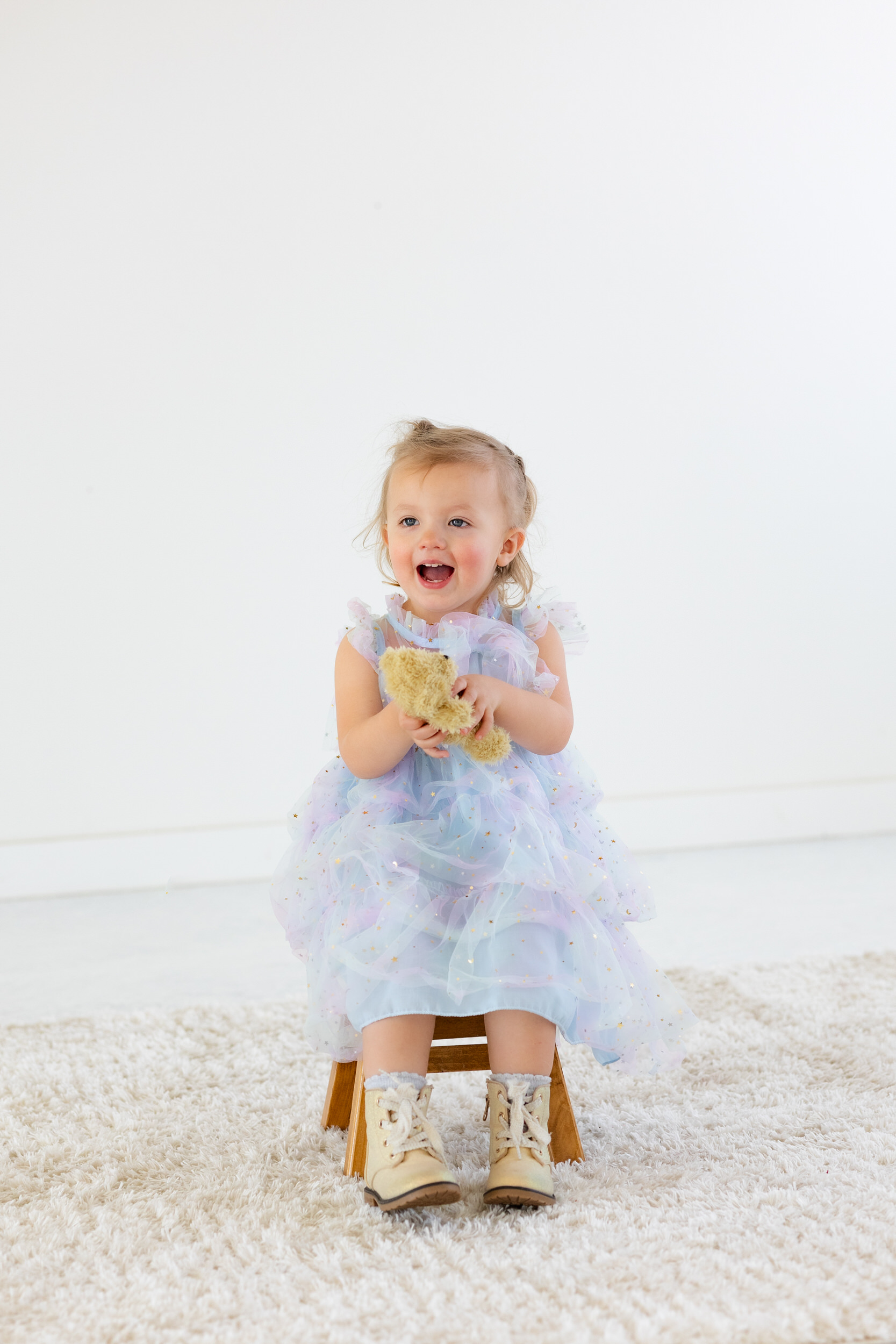 A two-year-old girl sits on a stool and holds a teddy bear.