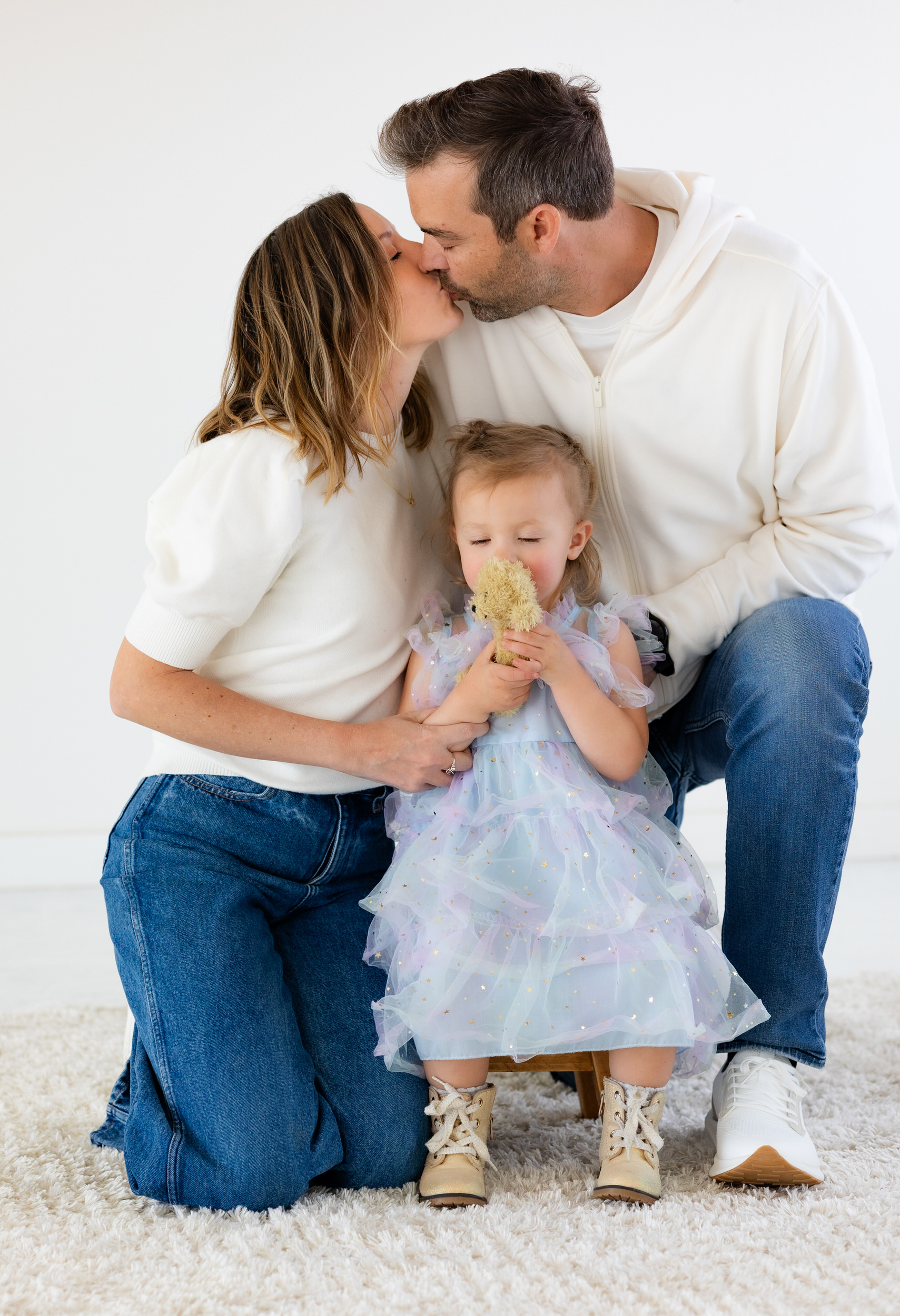 A mom and dad share a kiss while their two-year-old daughter kisses her teddy bear in front of them.
