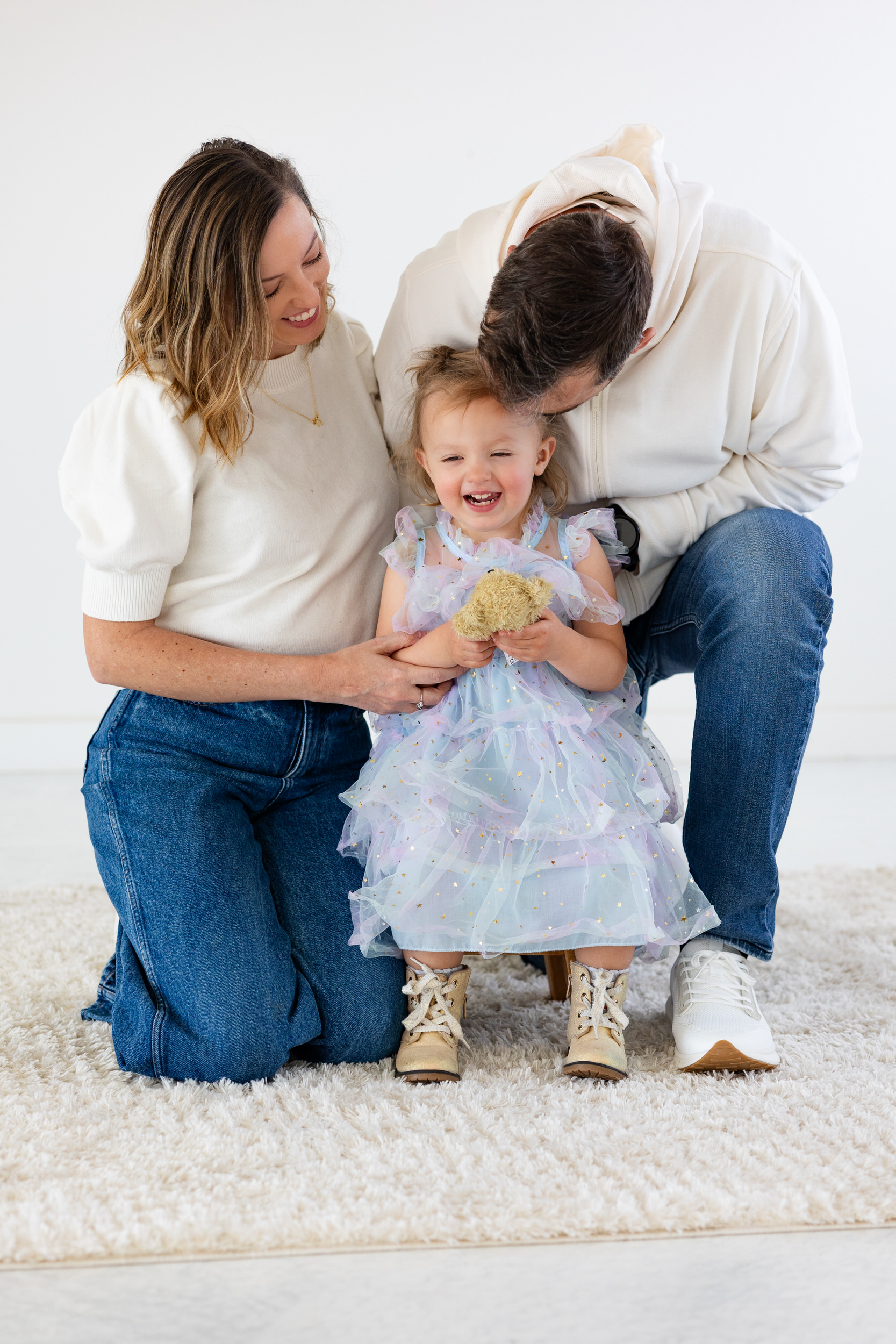 A mom smiles at her toddler daughter while her husband kisses their daughter on top of her head.