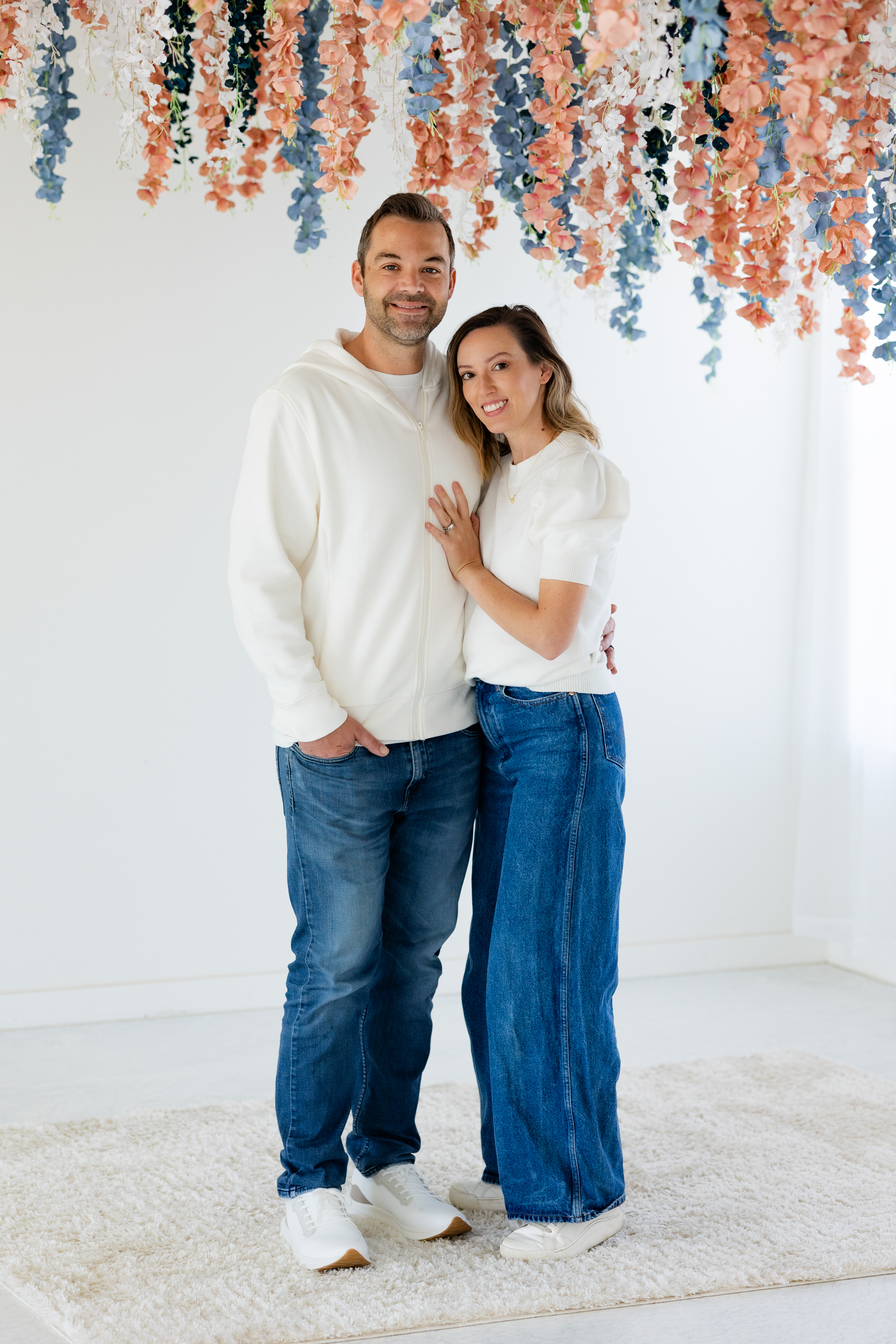 A wife leans into her husband with her hand on his chest and they both smile at the camera.