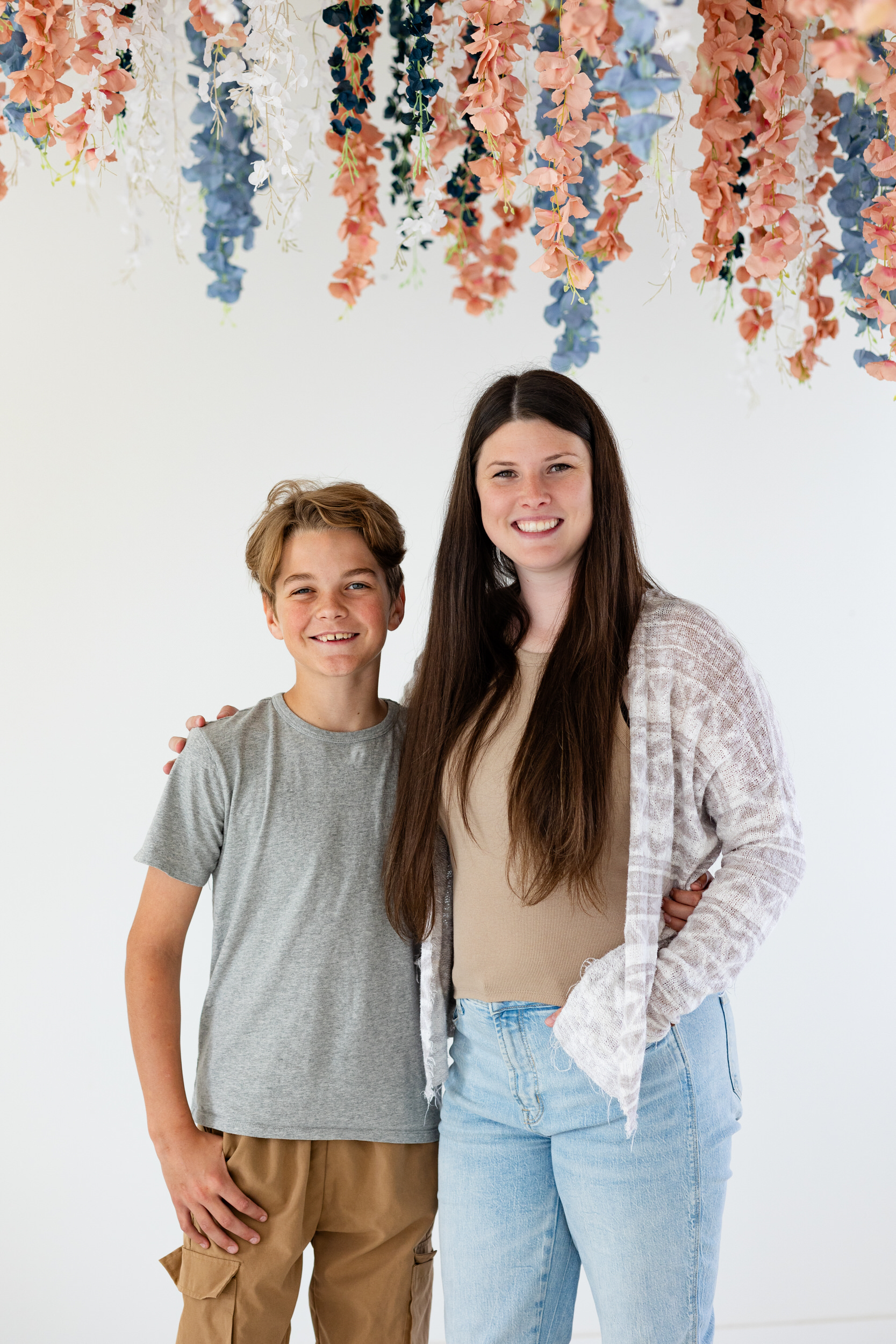 A mom and her pre-teen son stand next to each other with their arms around each other's shoulders and smile at the camera.