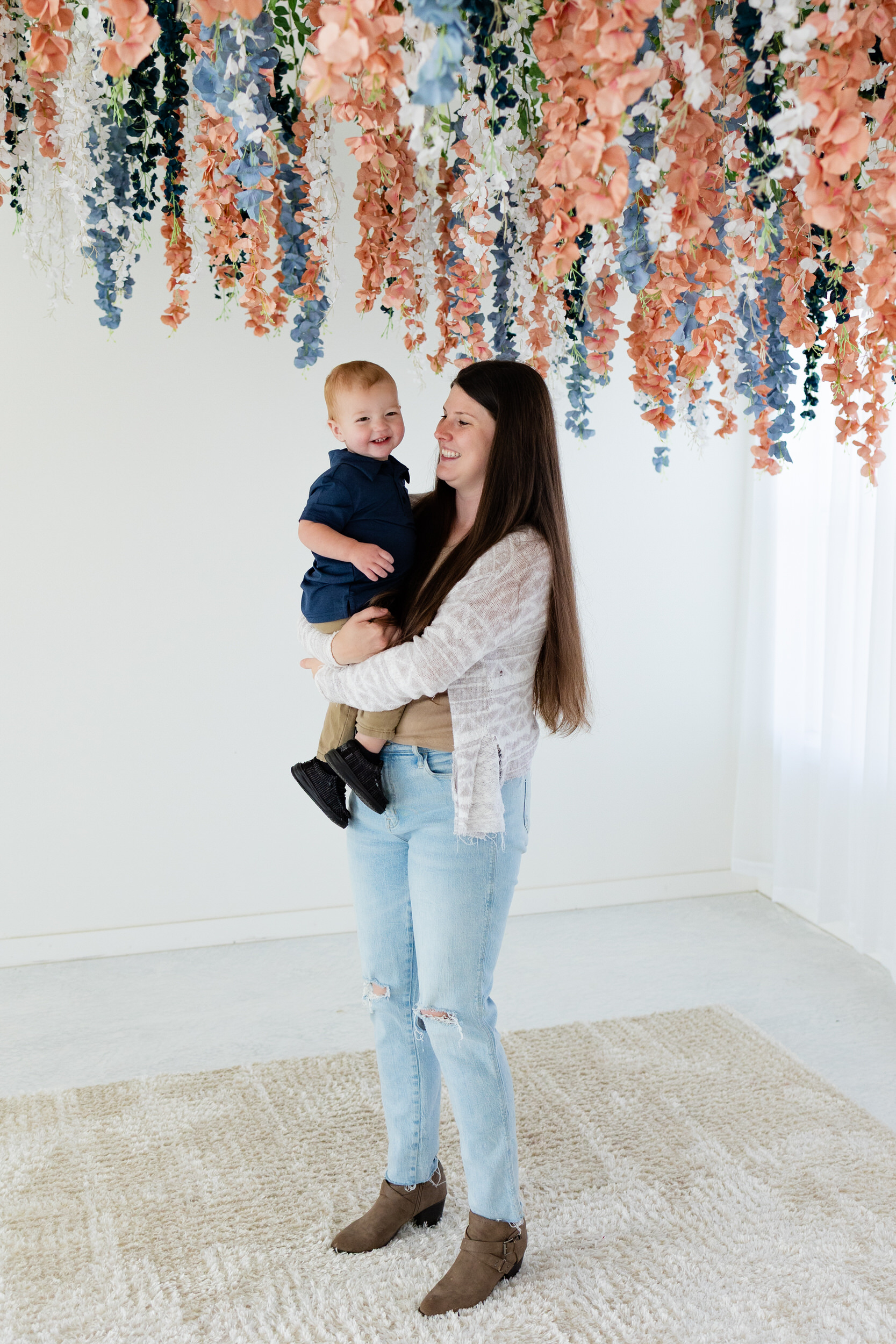 A mom holds her one-year-old son and smiles at him while he smiles at the camera.