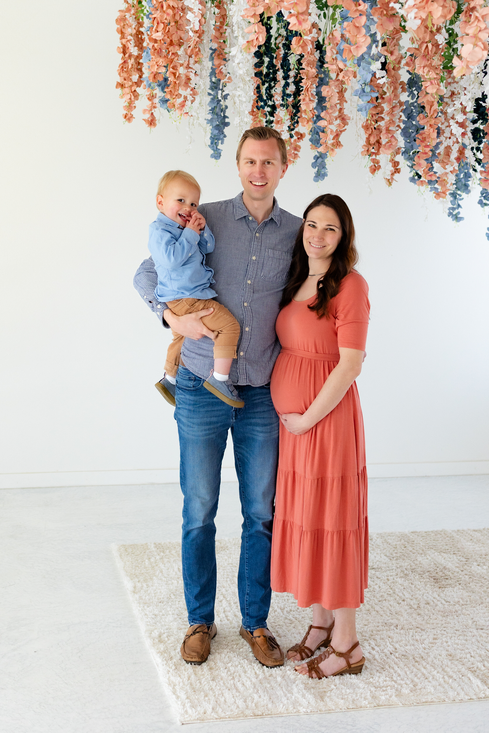A mom and dad hold their toddler son and smile at the camera while mom has a hand on her pregnant belly.