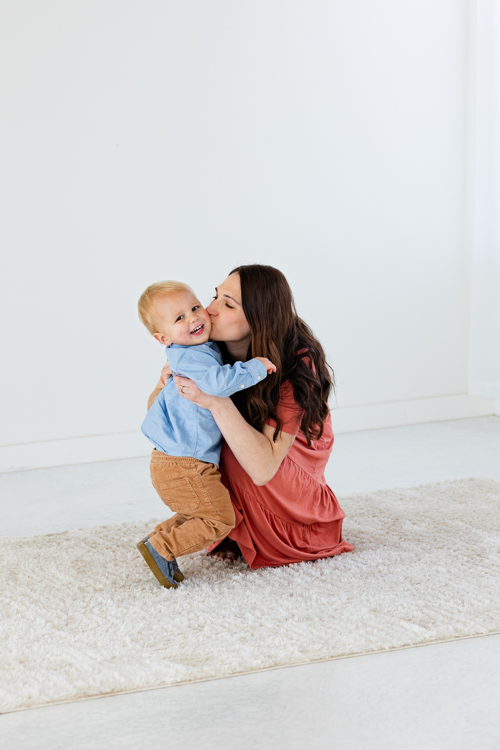 A mom squats down and hugs her toddler son and kisses him on the cheek while he smiles.