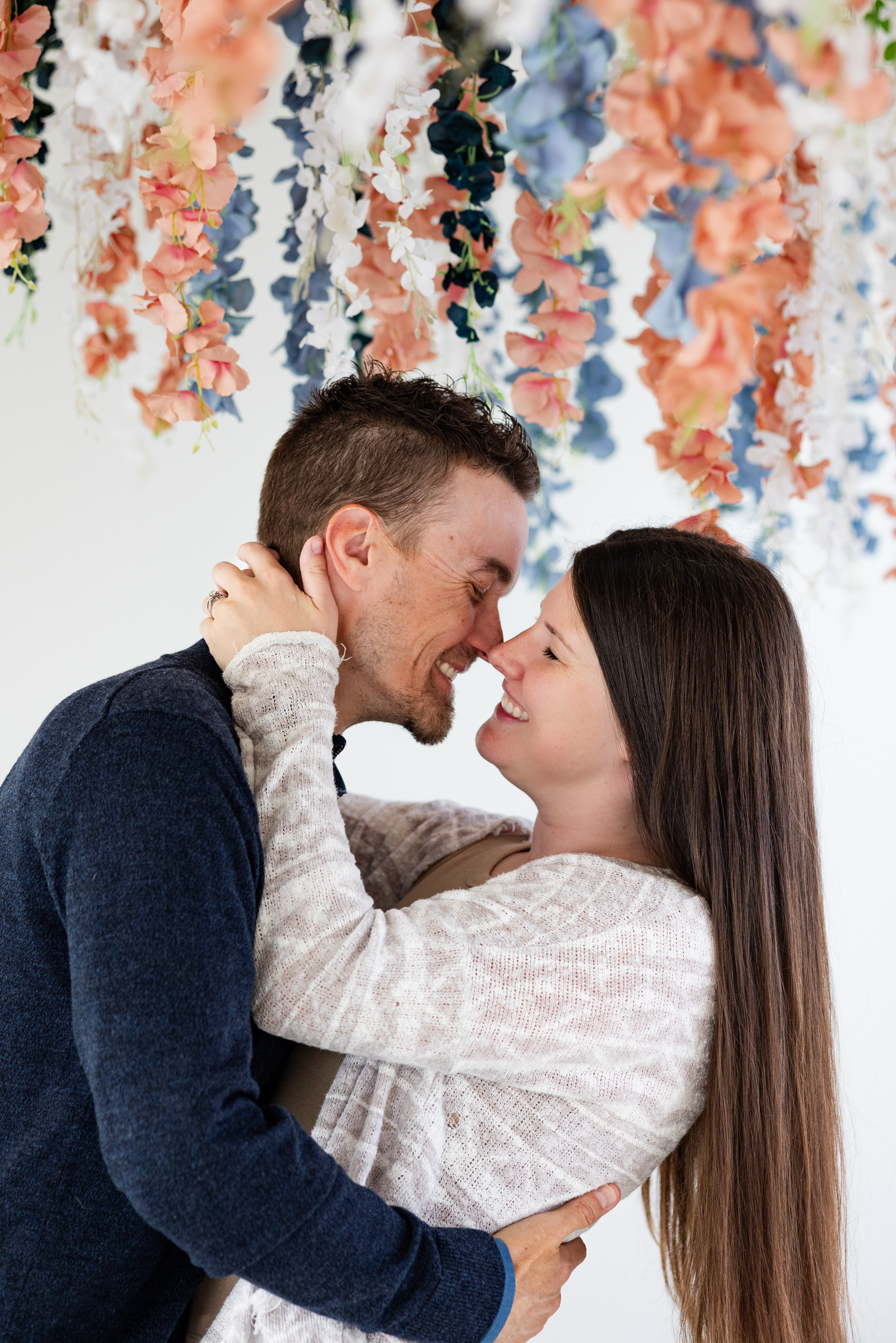 A husband and wife stand nose to nose and smile at each other.