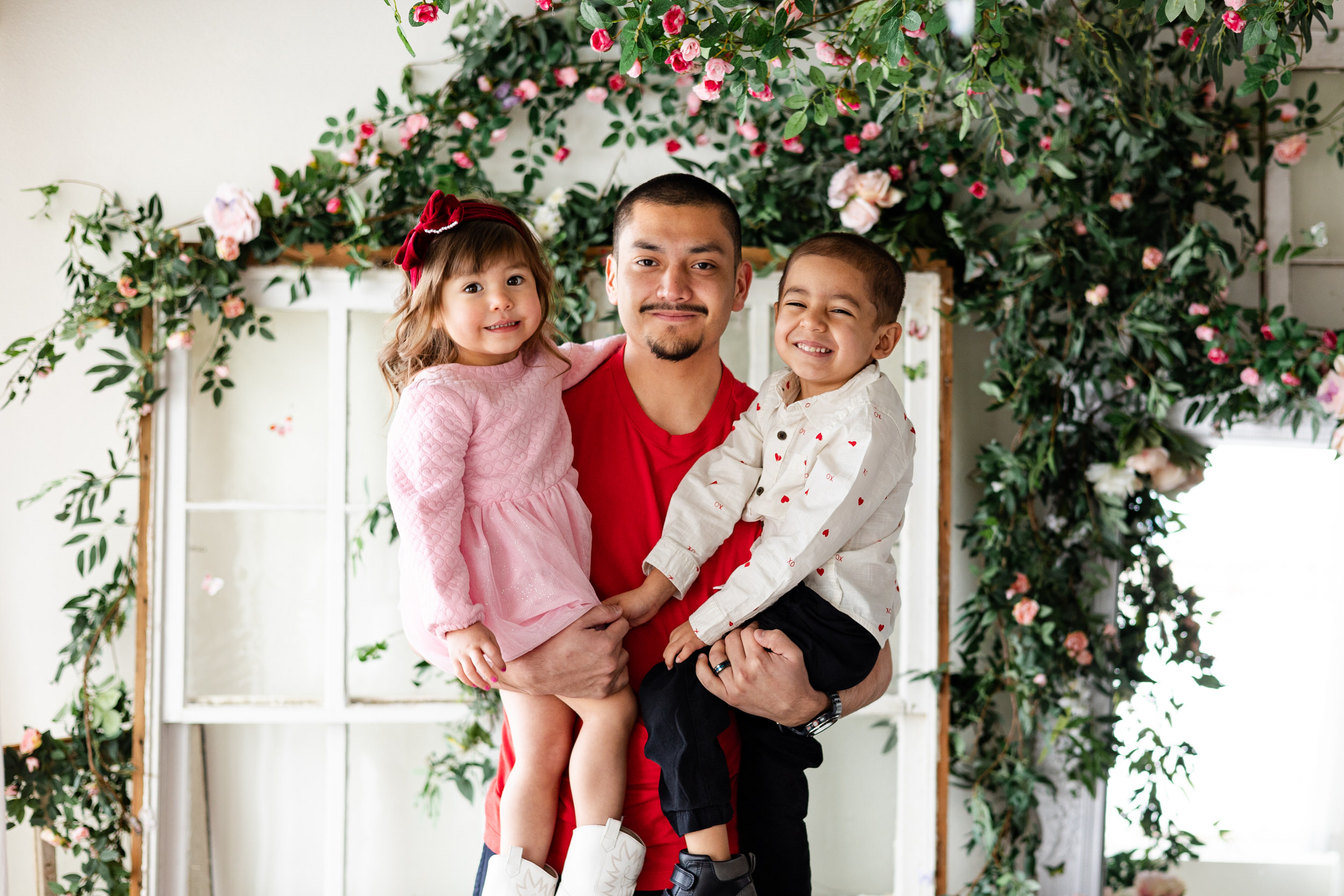 A dad holds his young son and daughter on his hips and they all smile at the camera.