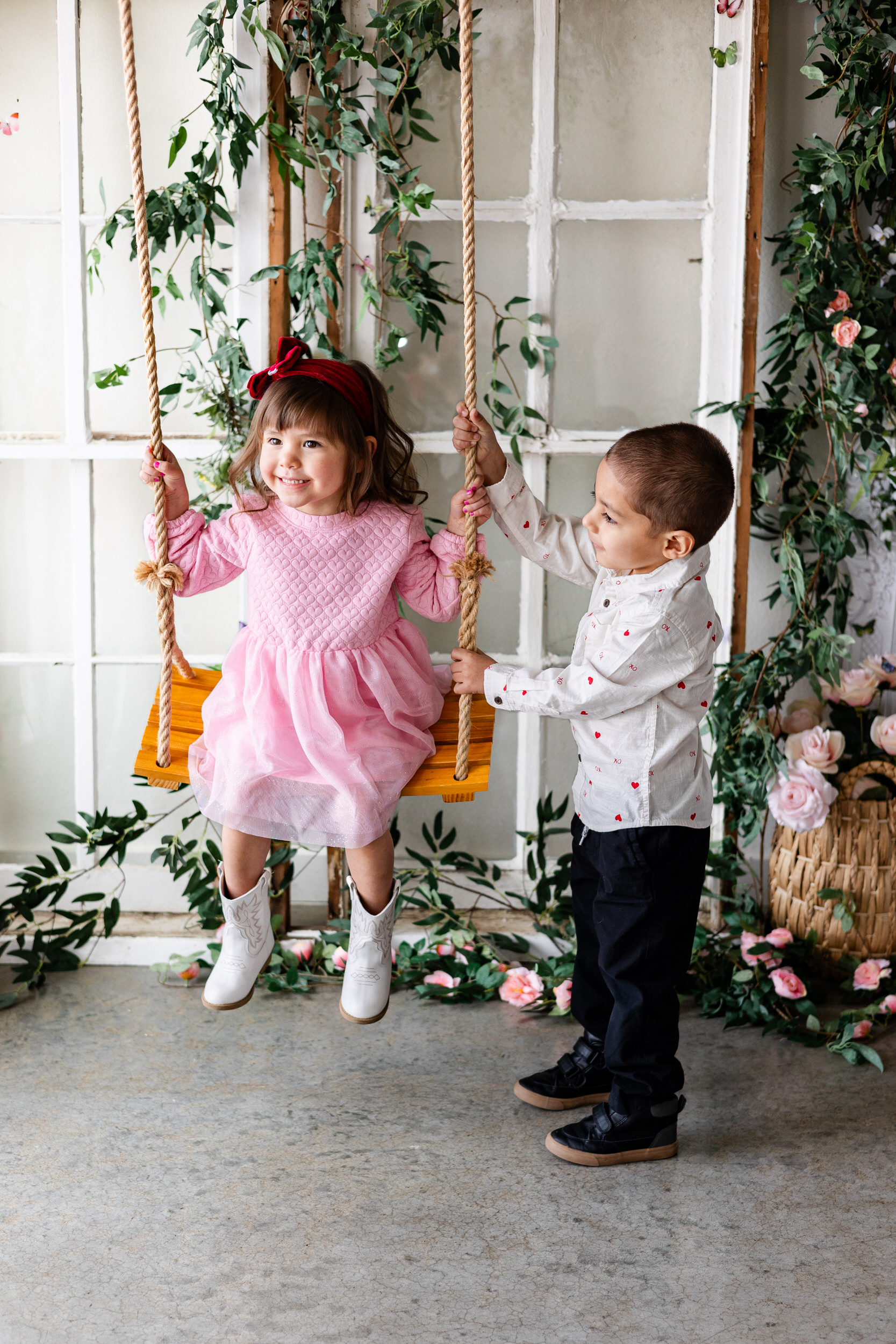 A little girl sits on a wooden swing and her older brother helps push her.