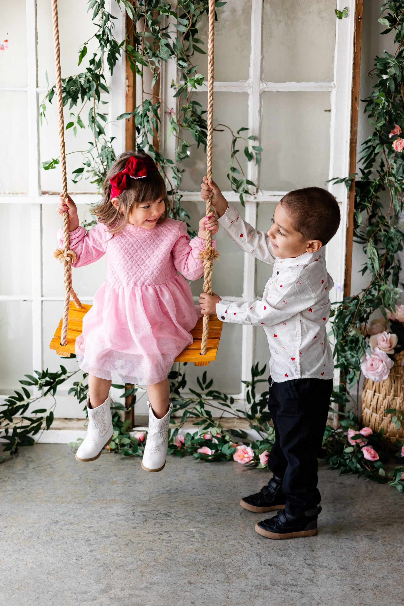 A little girl sits on a wooden swing and smiles at her older brother who helps push her.