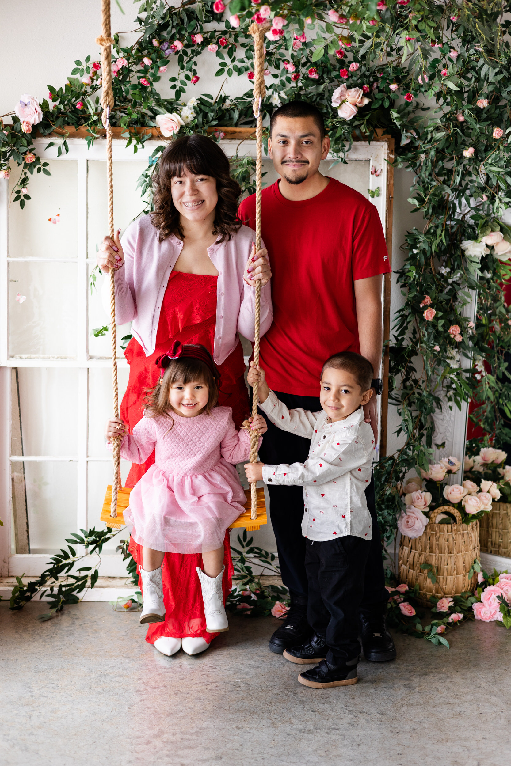 A little girl sits on a wooden swing and her mom, dad, and older brother gather around her and they all smile at the camera.