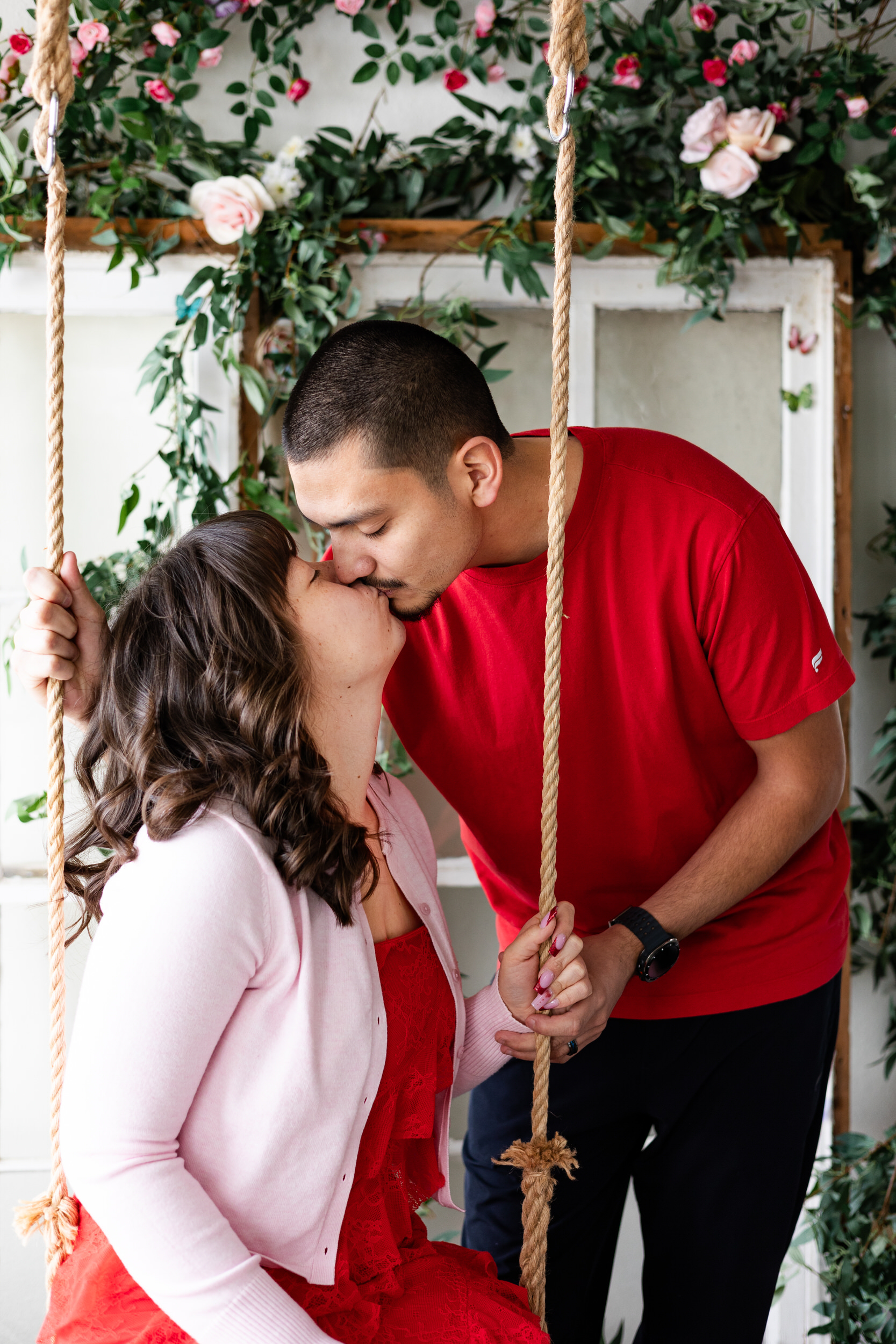 A woman sits on a wooden swing and her husband stands behind her and leans down to kiss her.